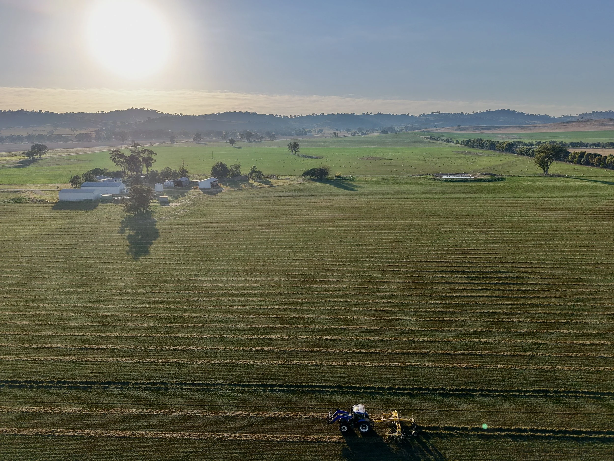 An aerial view of a farm field with a tractor working the land, several buildings in the background, and the sun shining in the sky.