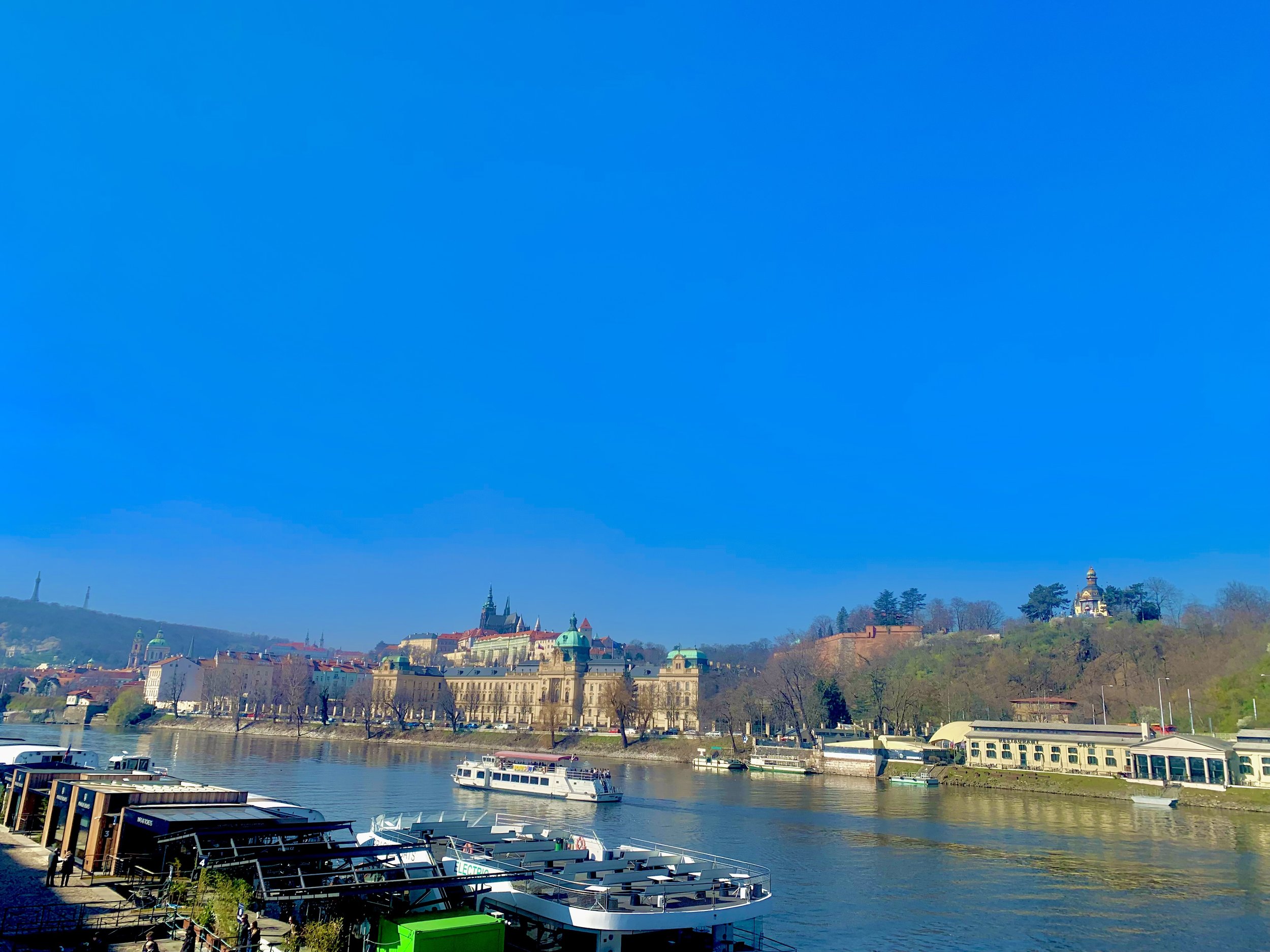 View of a river with boats, historic buildings along the riverbank, and a hill with trees and a small structure on top, under a clear blue sky.