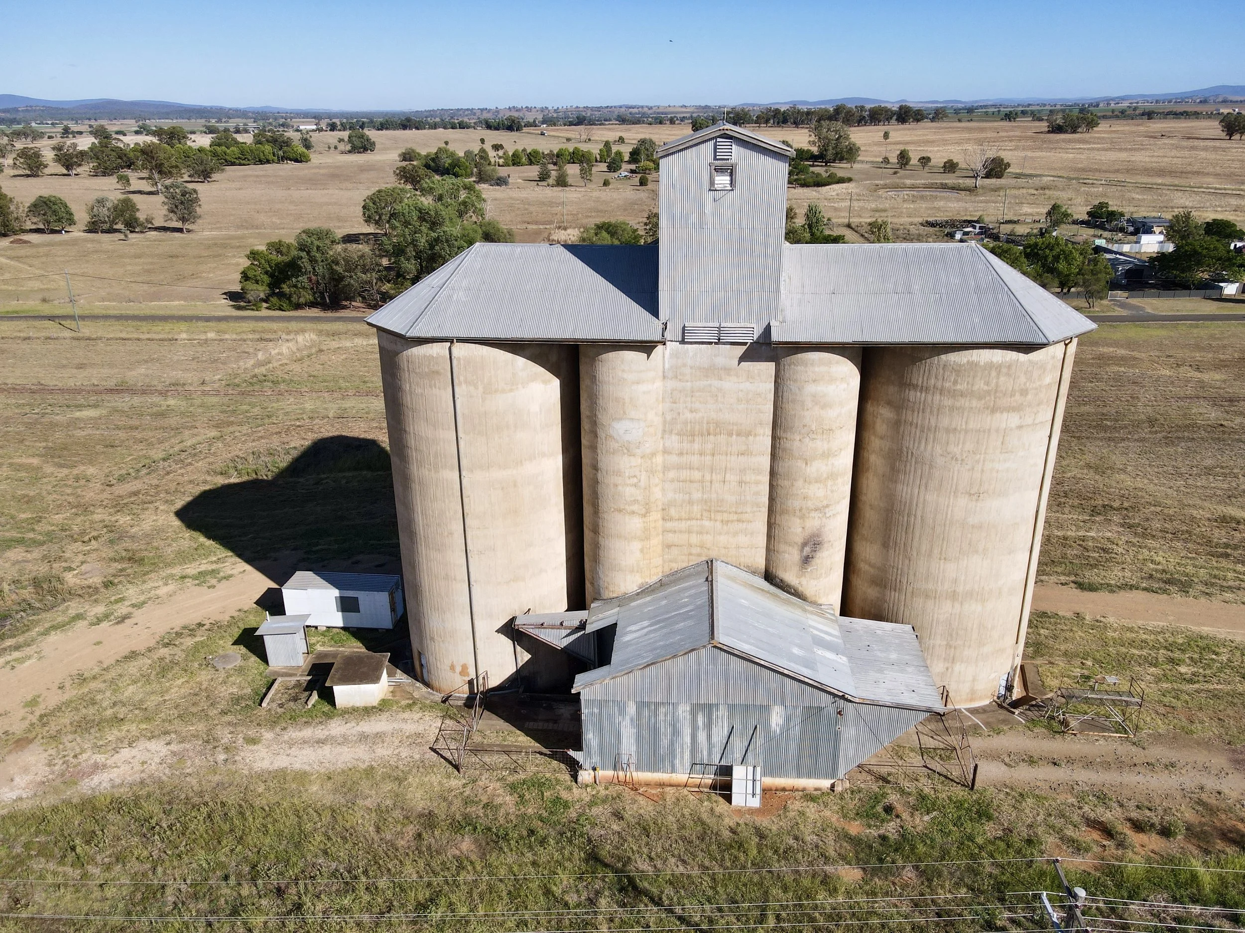 An old grain elevator with four large cylindrical silos, metal roofs, and a small adjacent metal building, situated in an expansive rural landscape with fields, trees, and distant hills.