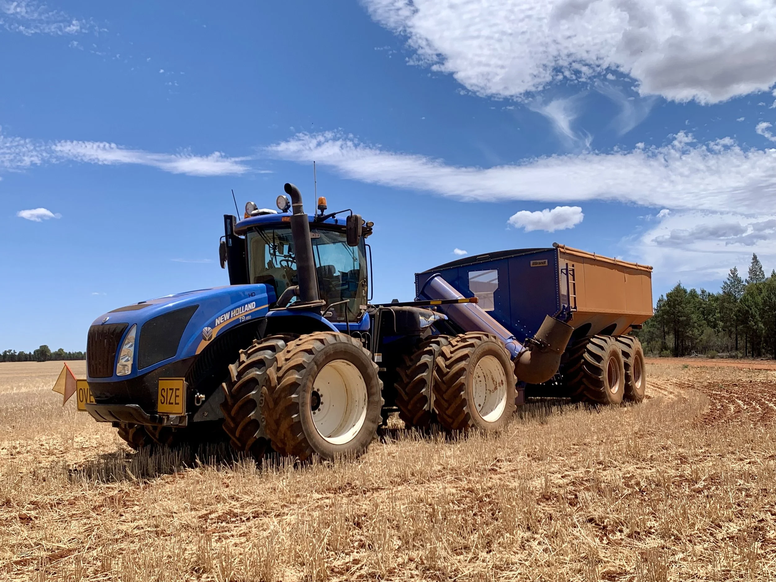A blue New Holland tractor pulling a large orange and blue agricultural implement in a dry field under a partly cloudy sky.