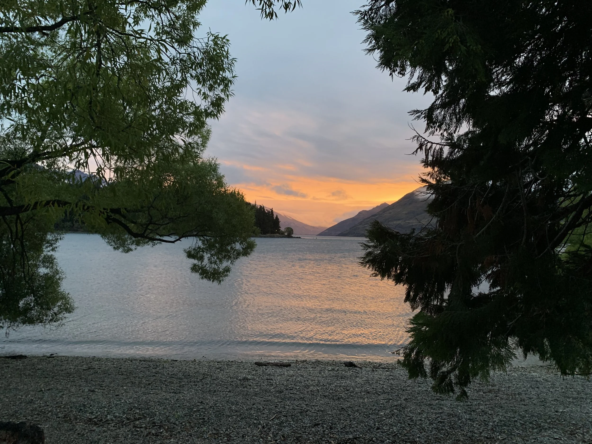 Sunset over a lake with mountains in the distance, framed by overhanging green trees on a rocky shore.