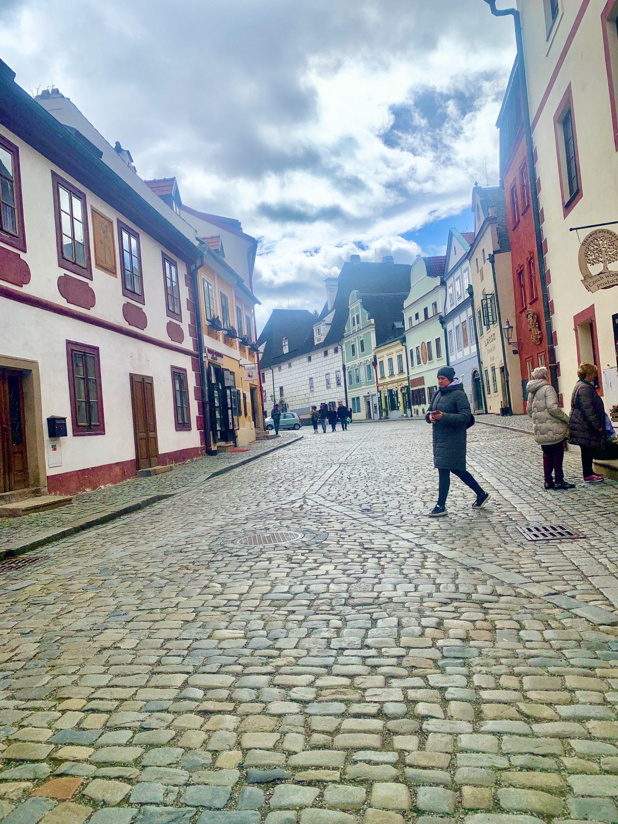 A cobblestone street lined with colorful European-style buildings on a partly cloudy day with a few people walking and standing around.