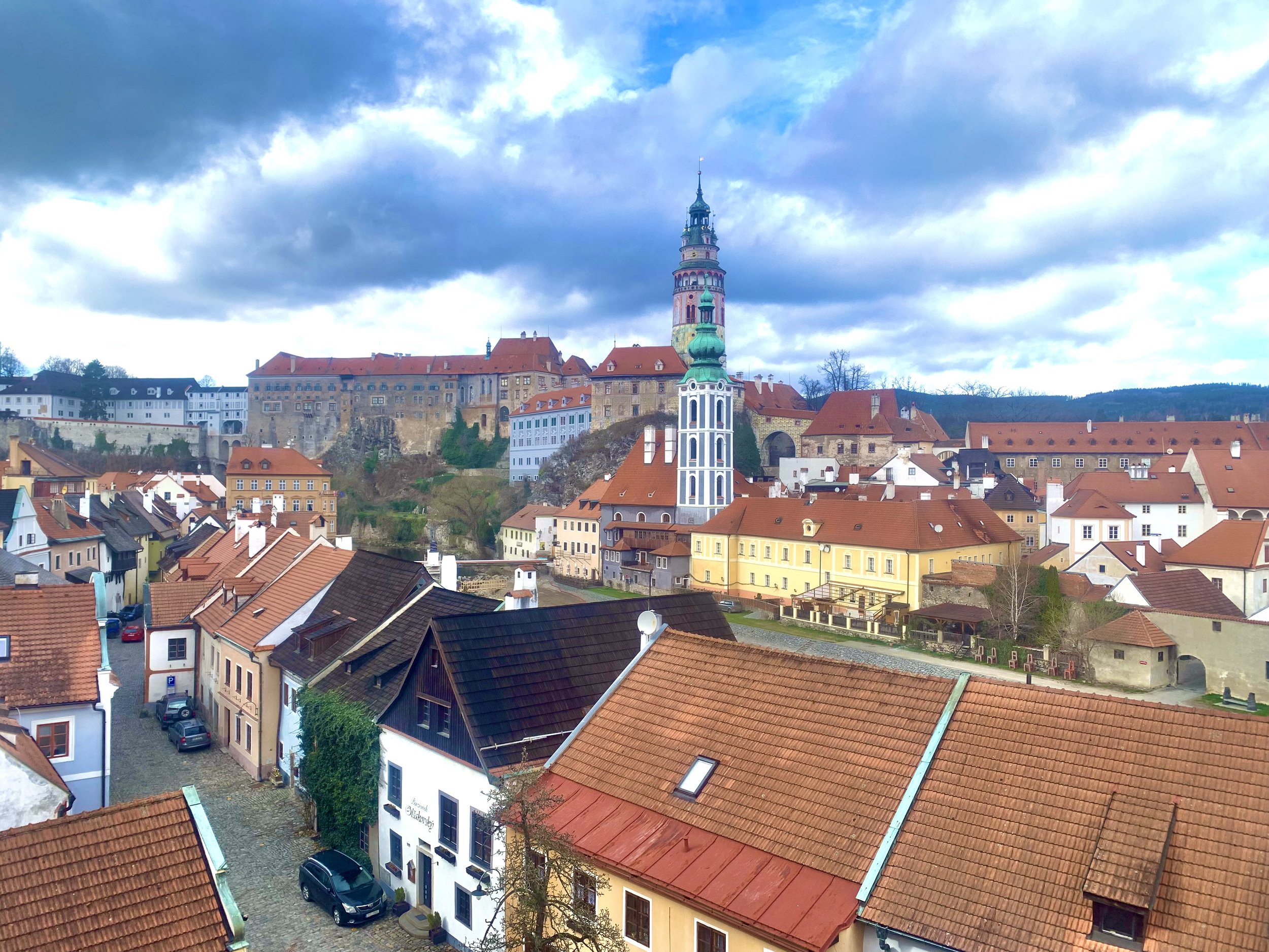 View of a European town with colorful buildings, including a tall church with a green dome and a spire, and a hilltop castle under a cloudy sky.
