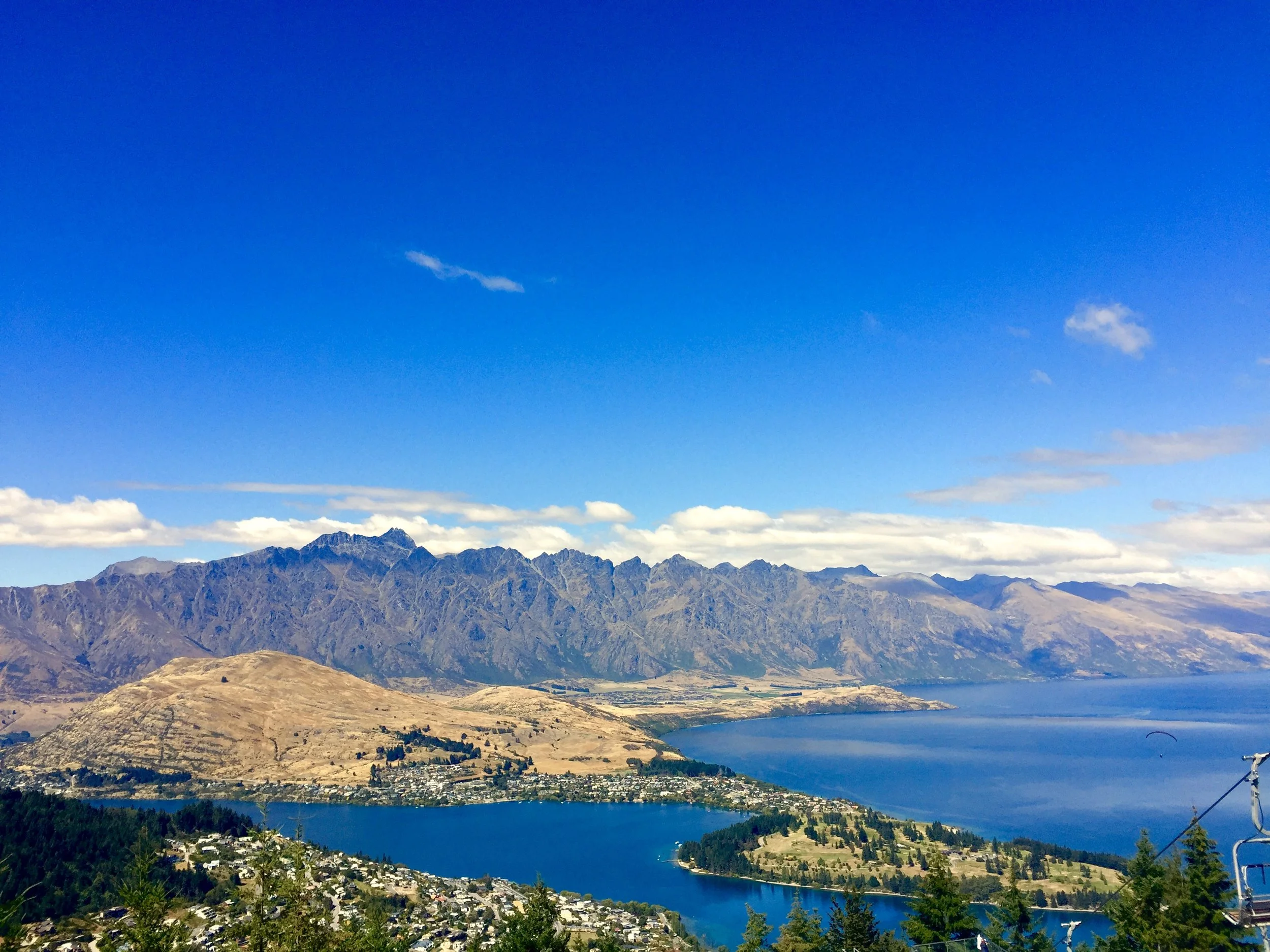 A scenic landscape featuring a large lake surrounded by hills and mountains under a bright blue sky with scattered clouds.