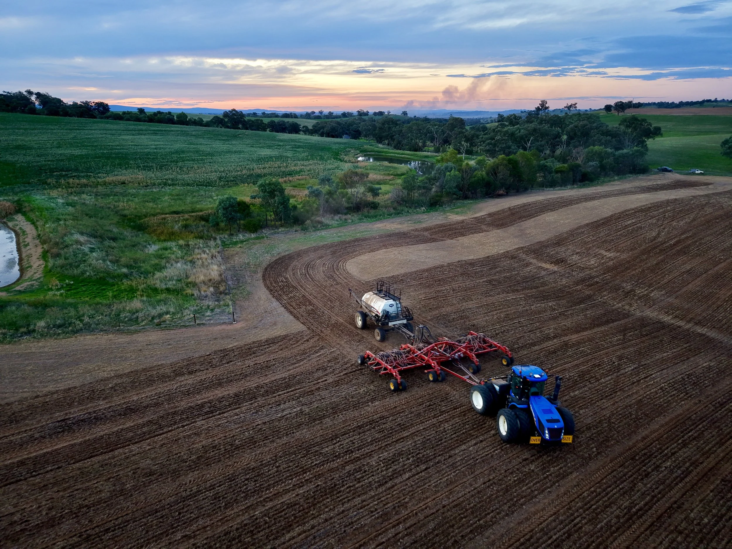 Aerial view of a large tractor planting crops on a field during sunset, with a green landscape and a sky filled with clouds.