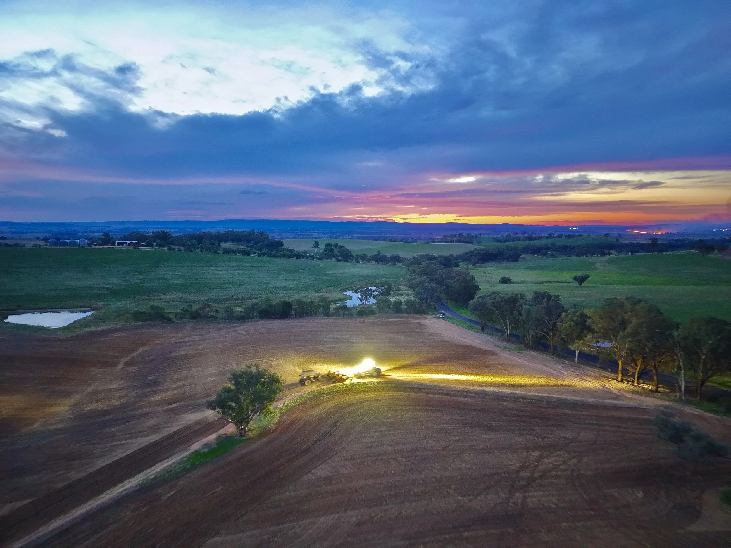 Aerial view of a countryside landscape at sunset with fields, trees, a small pond, and a dirt road.
