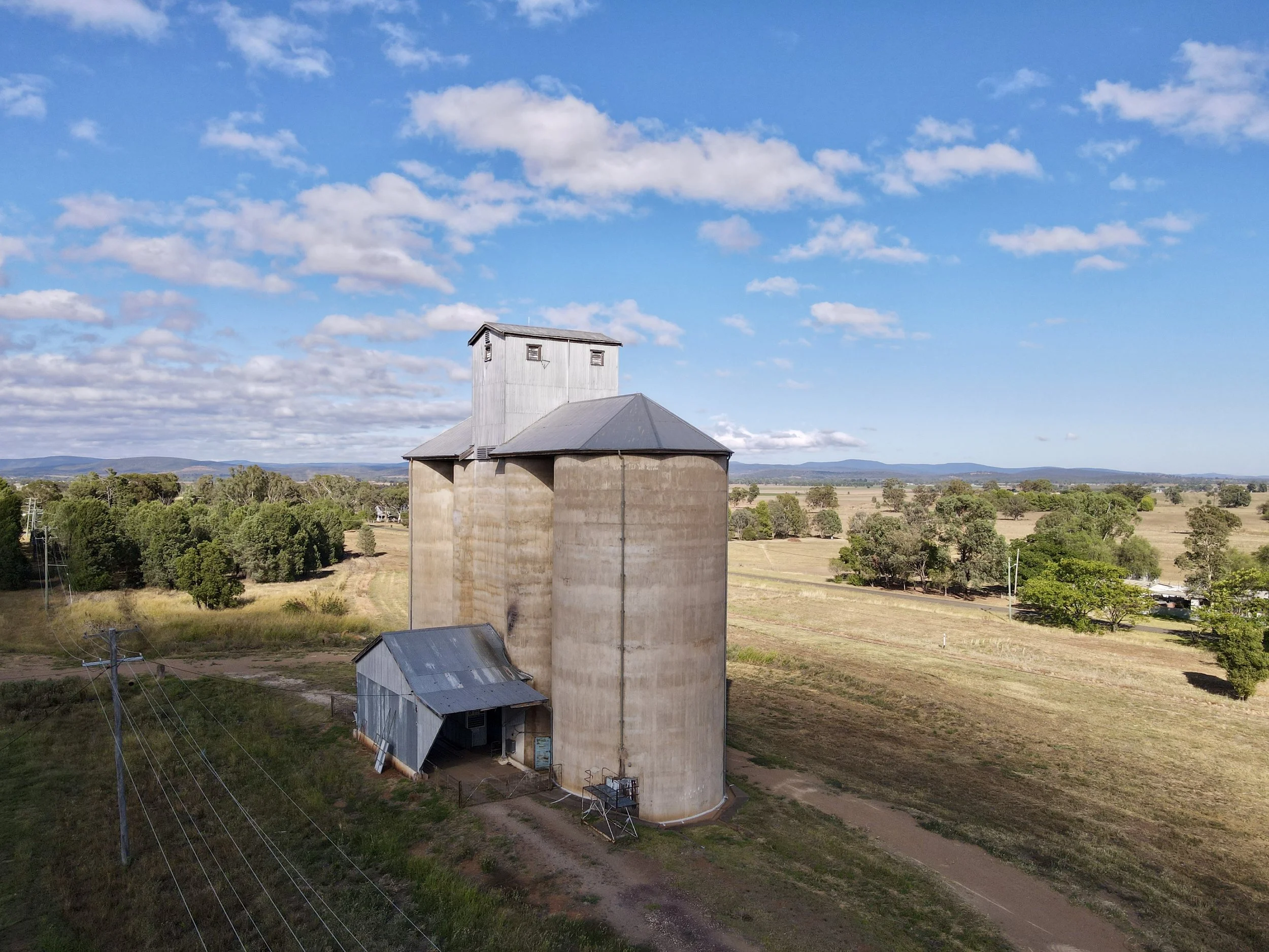 A rural landscape with a large concrete grain silo, a small metal shed, green trees, and open fields under a partly cloudy blue sky.