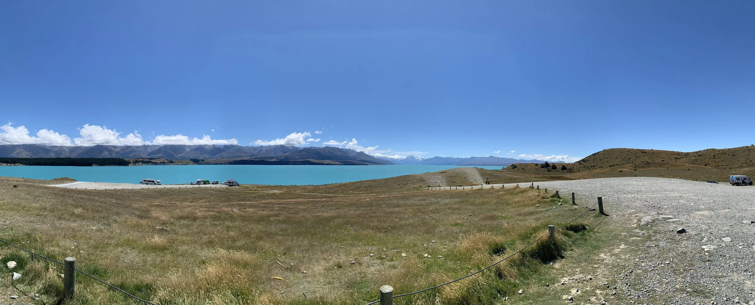 Scenic view of a turquoise lake surrounded by mountains, with a grassy area, a dirt parking lot, and several parked vehicles under a clear blue sky.