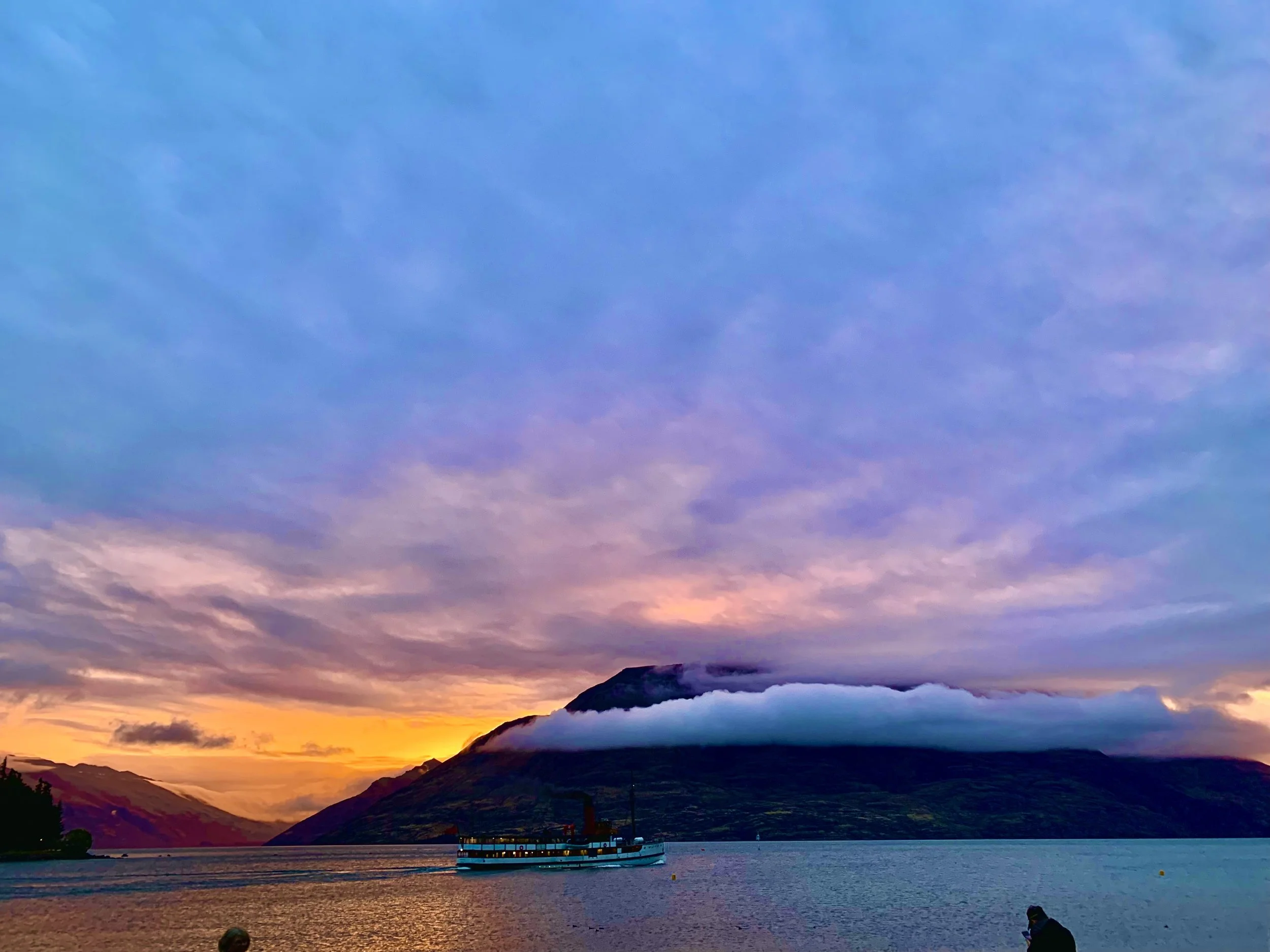 A vibrant sunset over a lake with mountains in the background, a boat on the water, and a person standing near the shore.