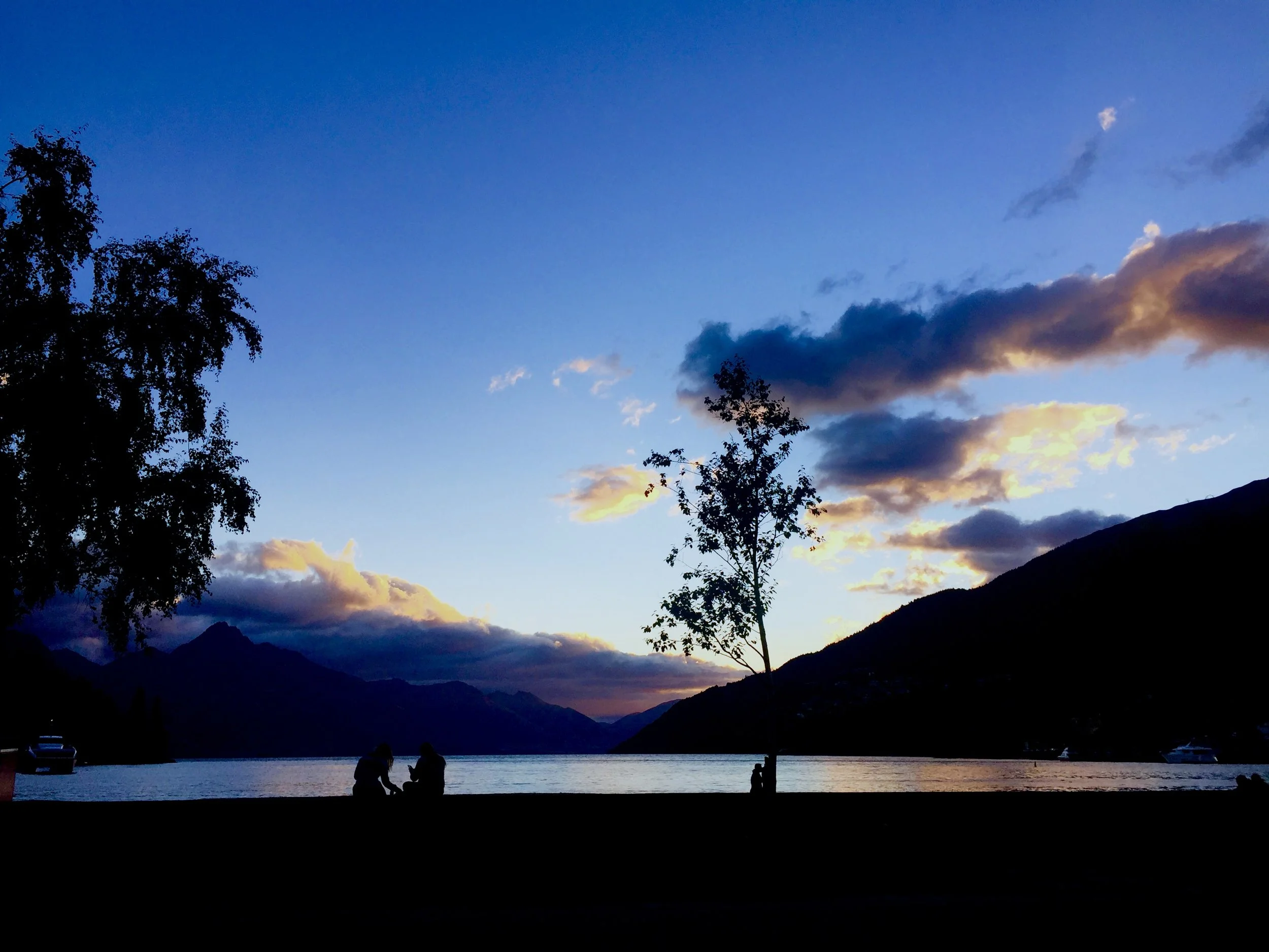 Silhouettes of people sitting beside a lake at sunset with mountains in the background and a partly cloudy sky.