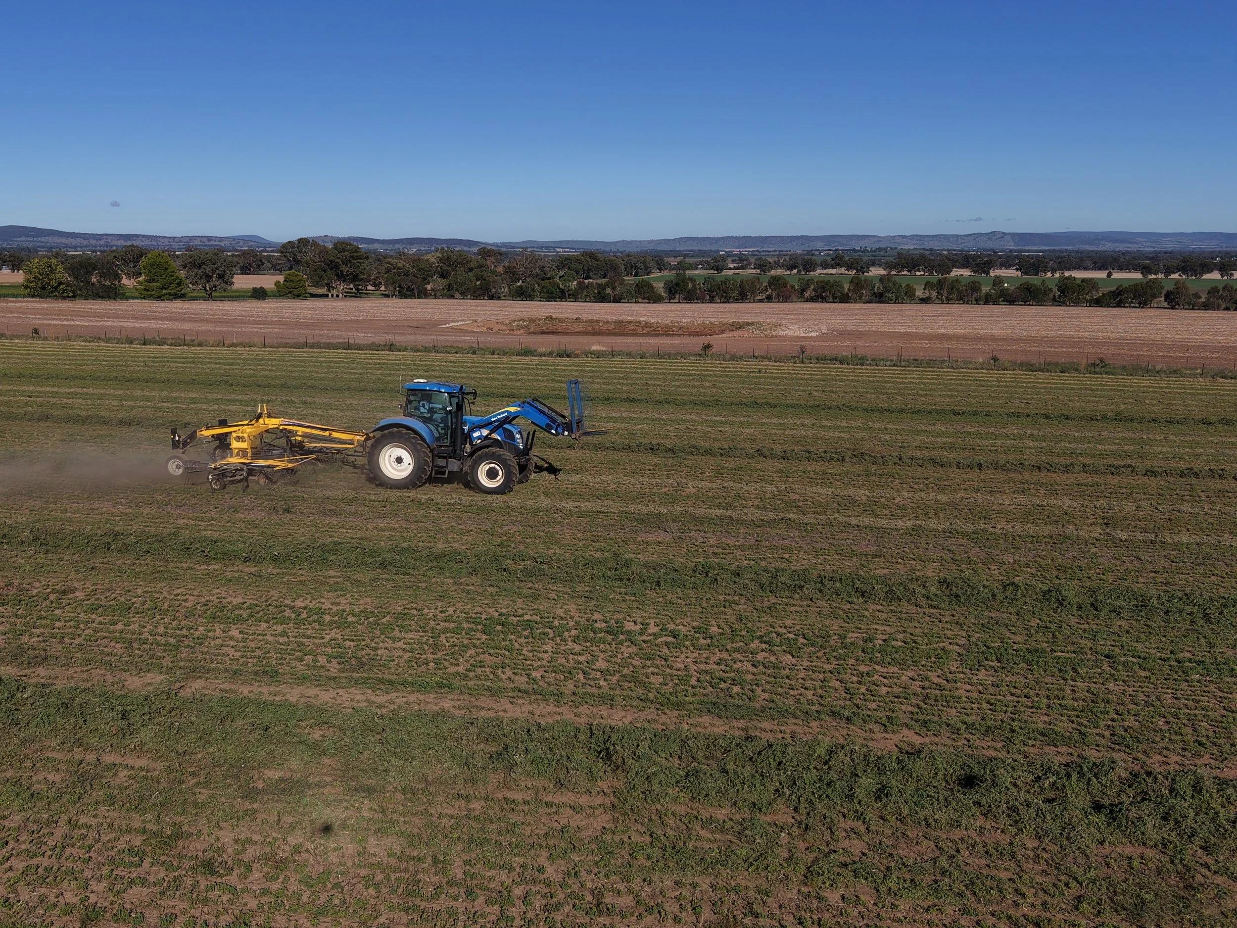 A blue tractor working on a field of young crops under a clear blue sky, with distant hills and farmland in the background.