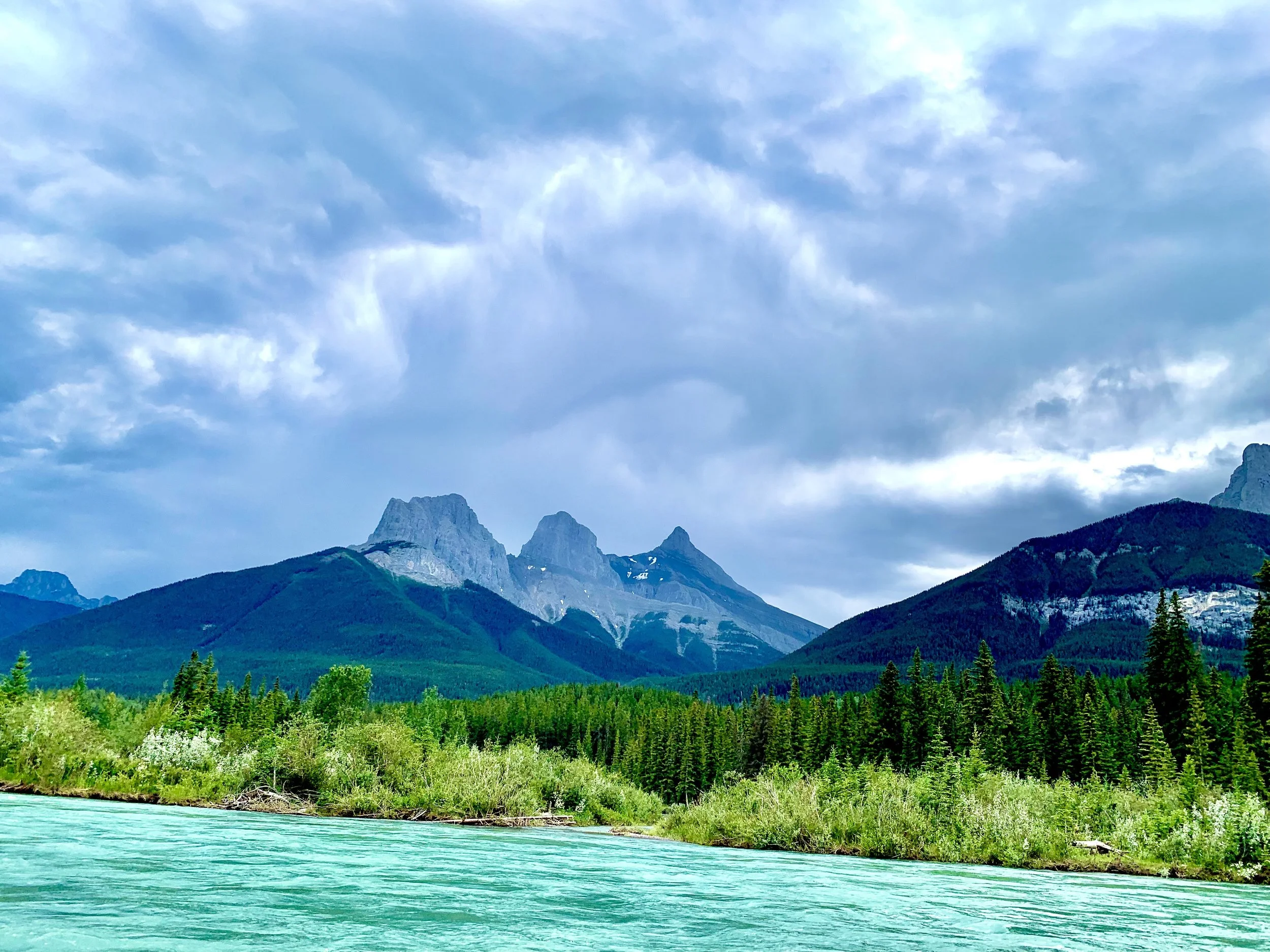 A mountain landscape with turquoise river in foreground, dense green forest, and rocky mountain peaks under a cloudy blue sky.