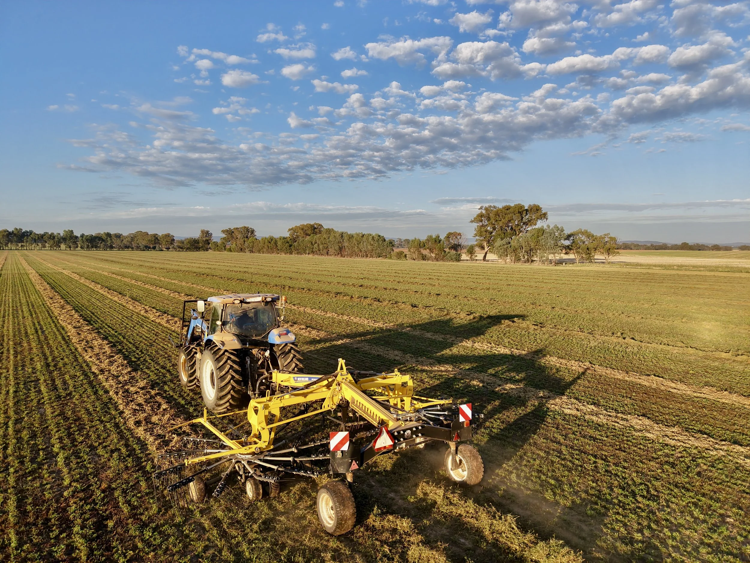 A tractor with a yellow implement attached is working on a large green field under a partly cloudy sky with some trees in the distance.