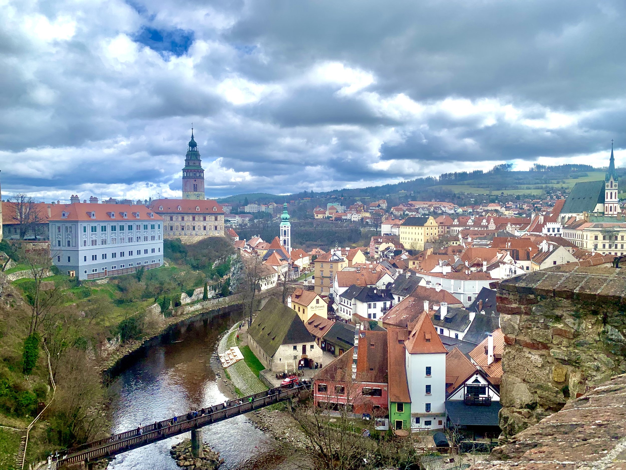 A scenic view of a historic European town with red-tiled roofs, a river with a pedestrian bridge, old buildings, and church steeples under a cloudy sky.