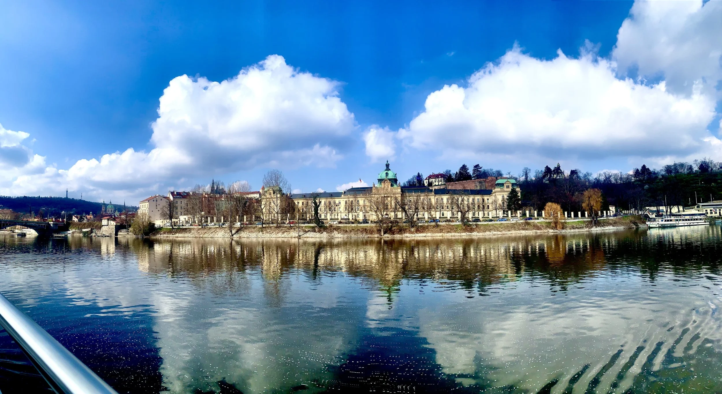 Historic building with a green dome on the bank of a river, with a partly cloudy sky reflected in the water.