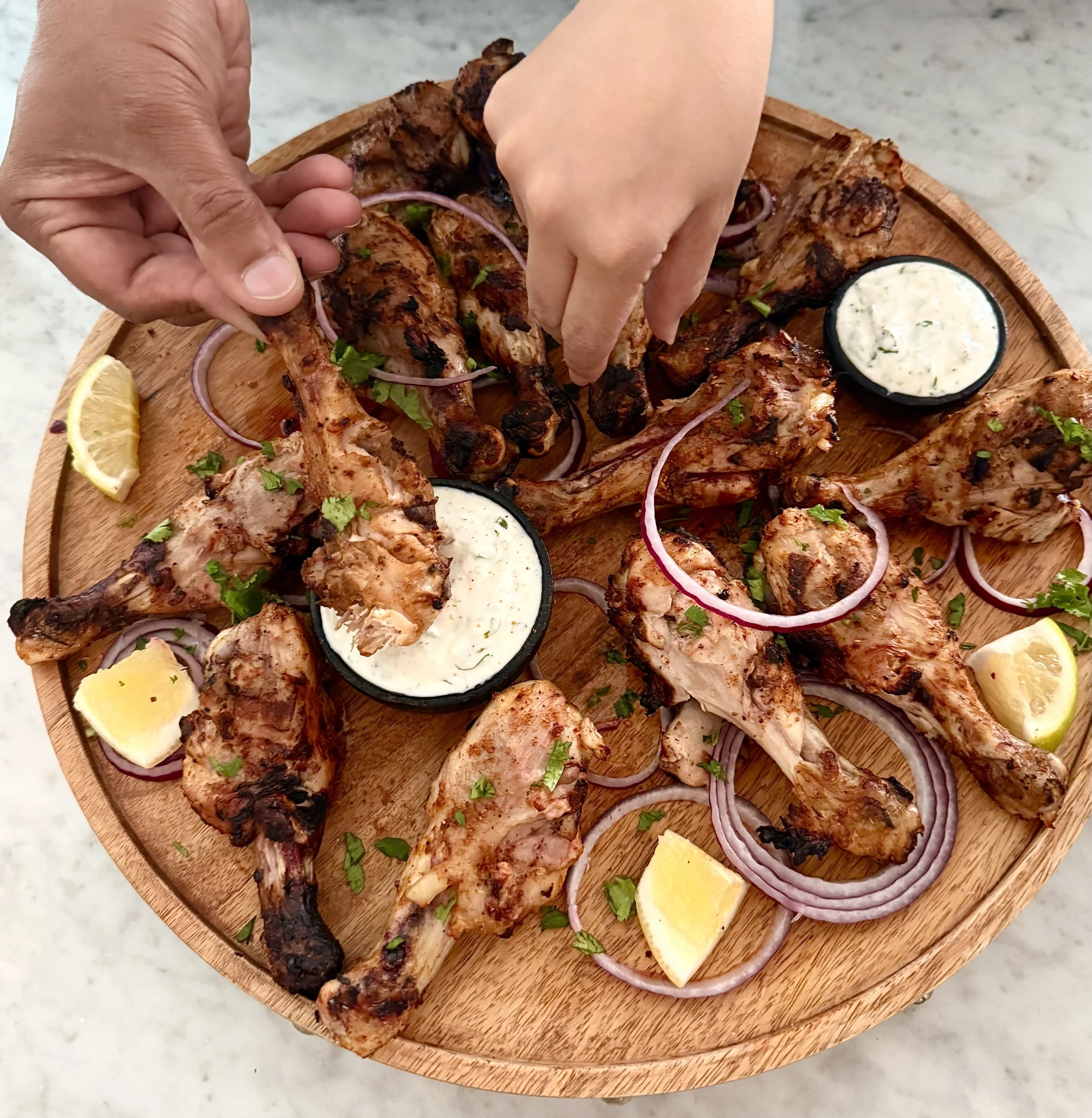 Tandoori chicken drumsticks on a wooden platter with yogurt sauce, red onion, cilantro and lemon wedges