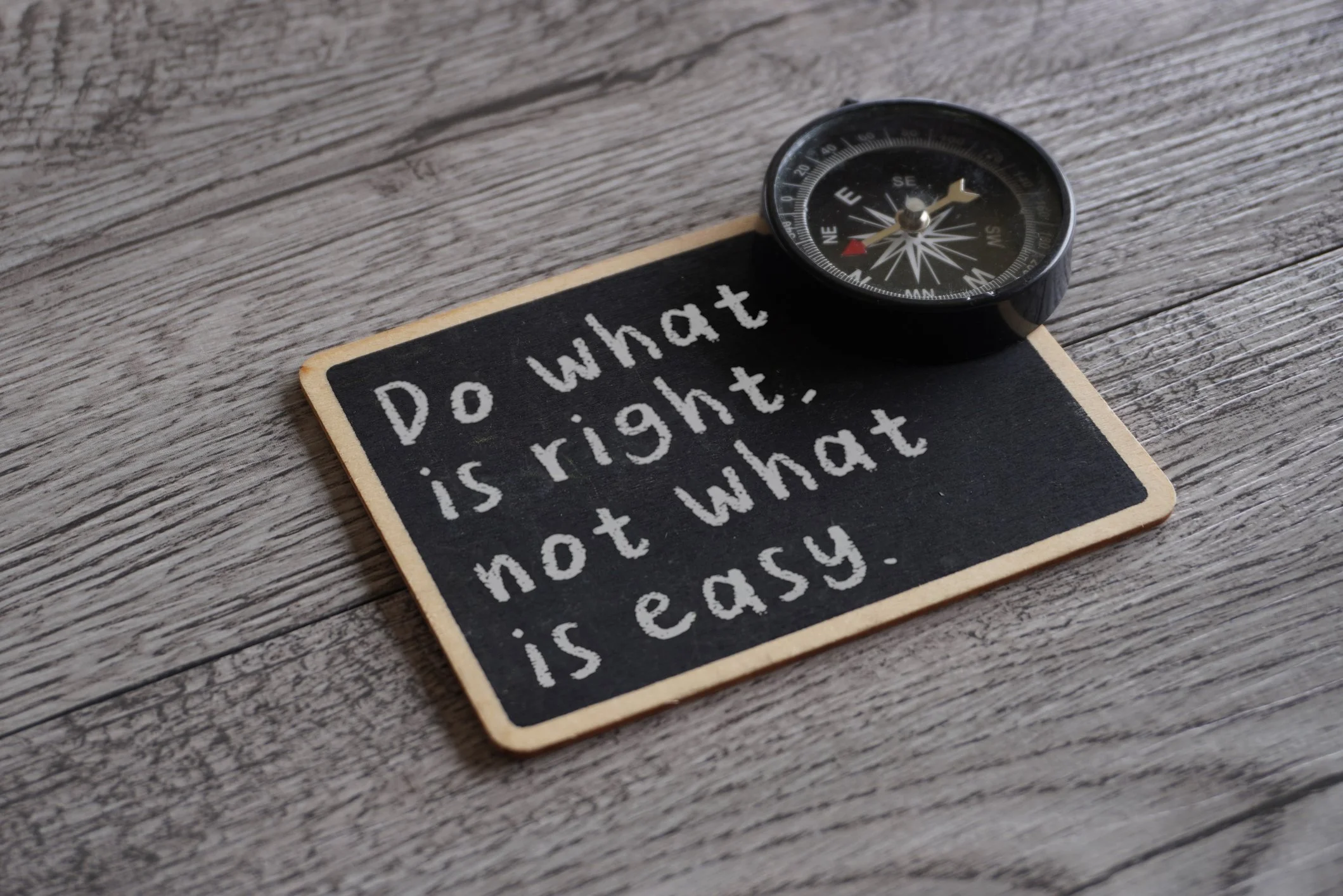 A small black chalkboard with a wooden border shows a motivational quote written in white chalk, saying 'Do what is right, not what is easy.' A black compass with a clear face is placed on the top right corner of the chalkboard, lying on a textured wooden surface.