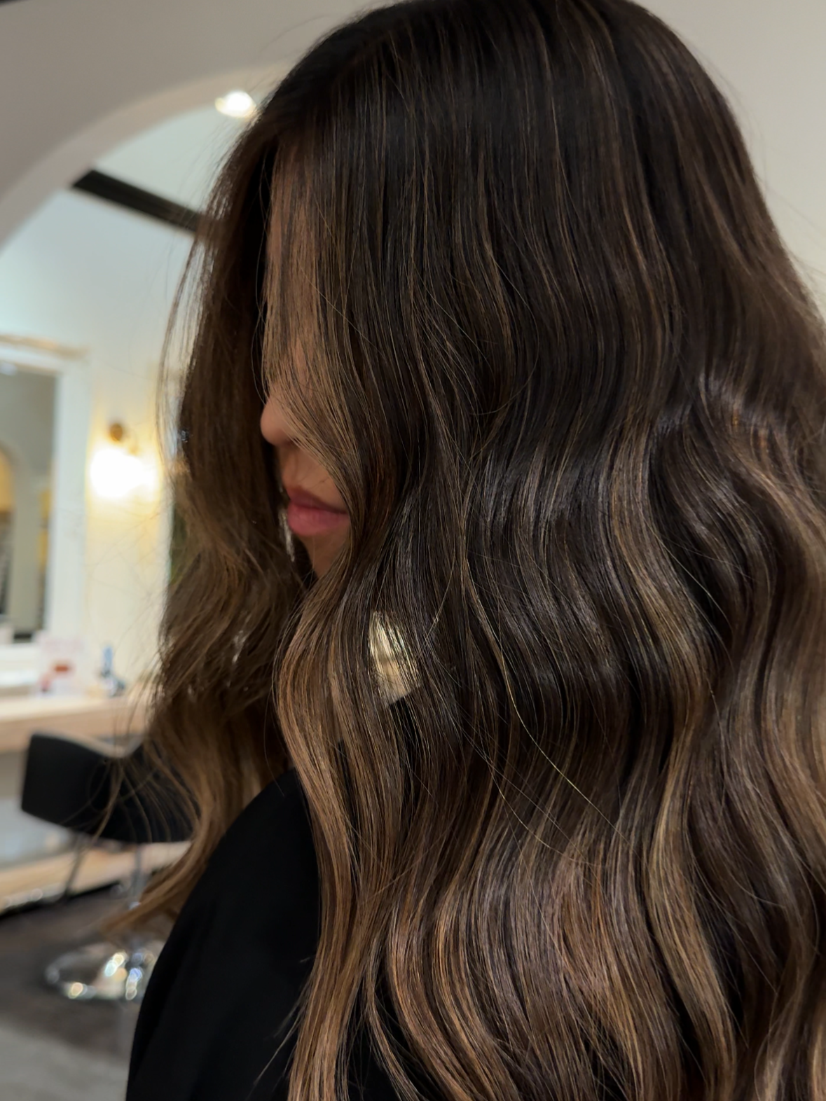 Close-up of a woman with long wavy brown hair with blonde highlights, looking downward, in a salon setting.
