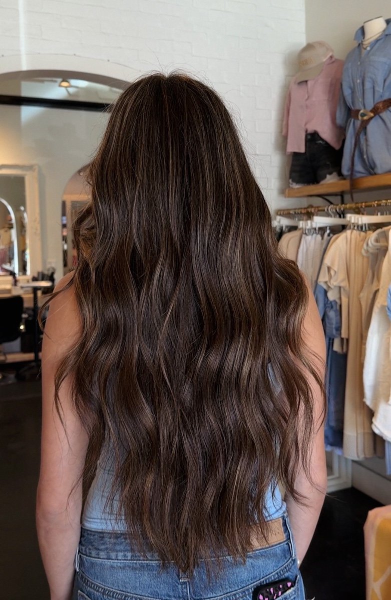 A woman with long, wavy brown hair inside a clothing store, with racks of clothes and mannequins in the background.