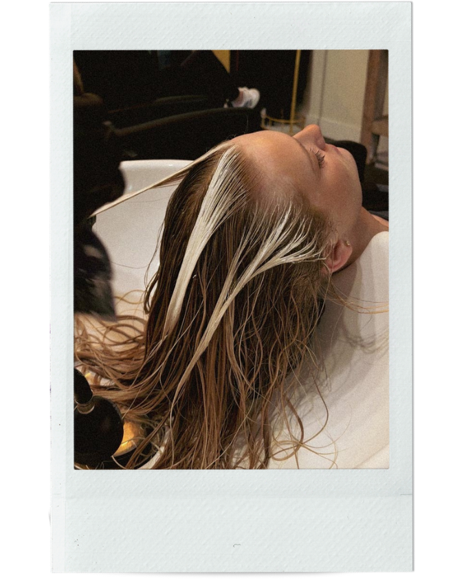 A woman with wet, blonde hair reclining with her head tilted back at a salon sink, receiving a hair wash.