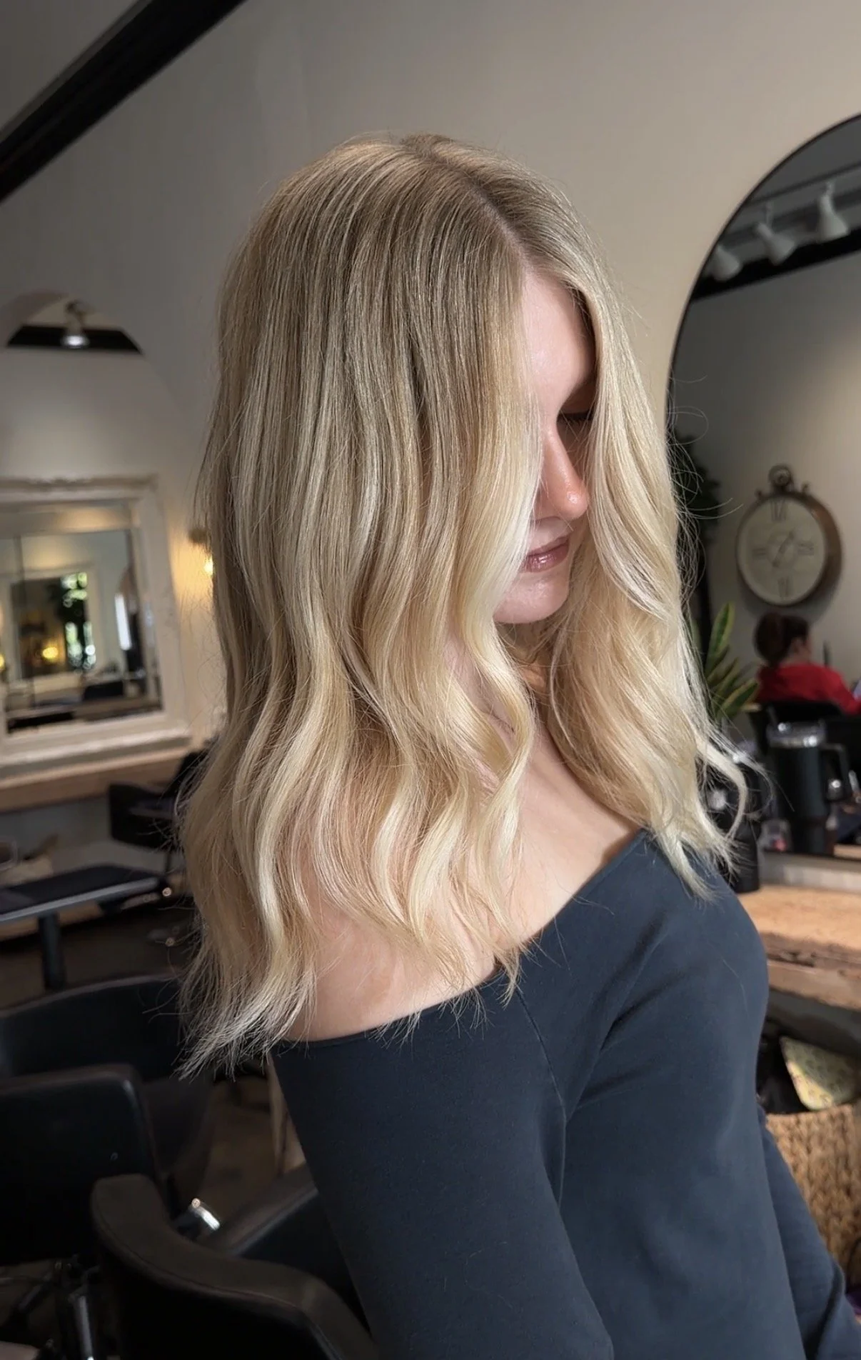 Woman with wavy blonde hair in a salon, wearing a black top, with salon mirrors and chairs in the background.