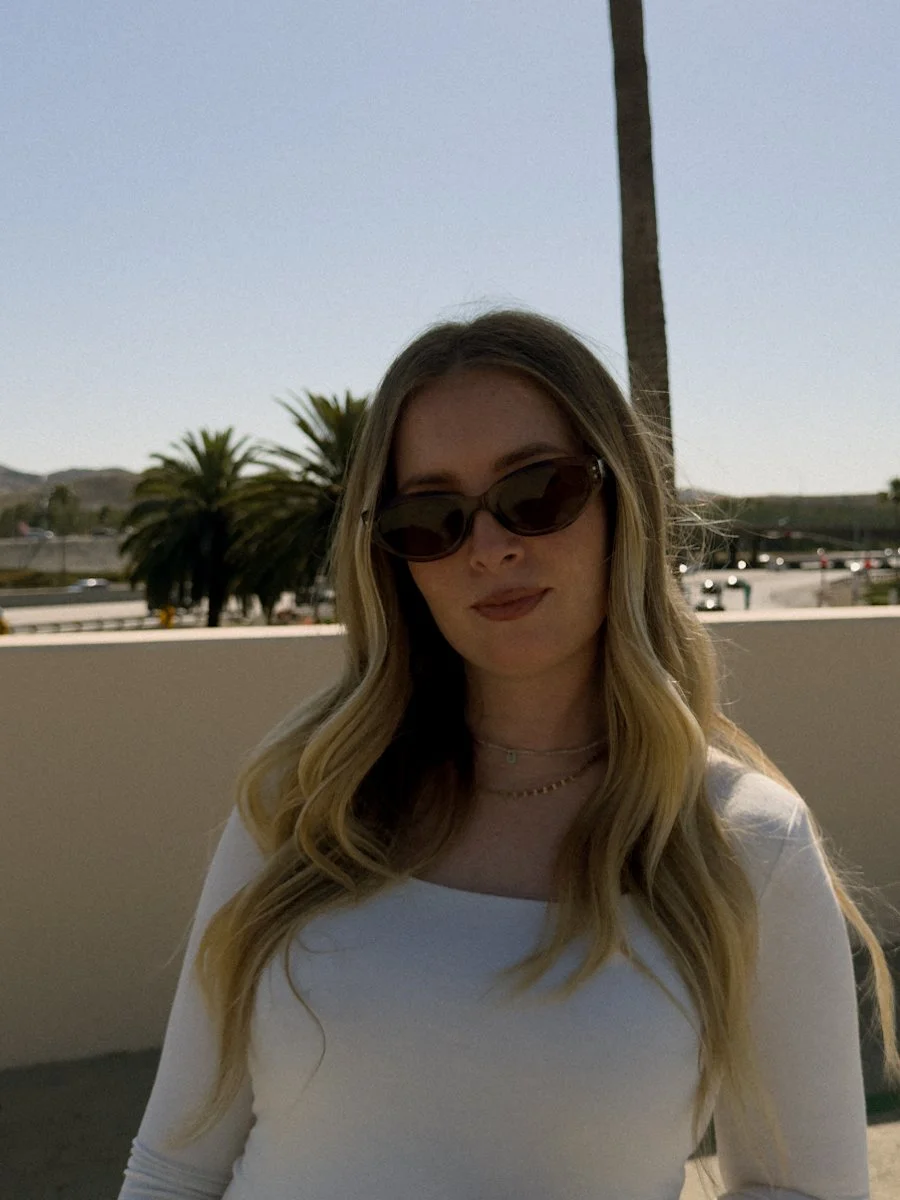 Young woman with long blonde hair, wearing sunglasses and a white top, standing outdoors near palm trees with a clear blue sky behind her.