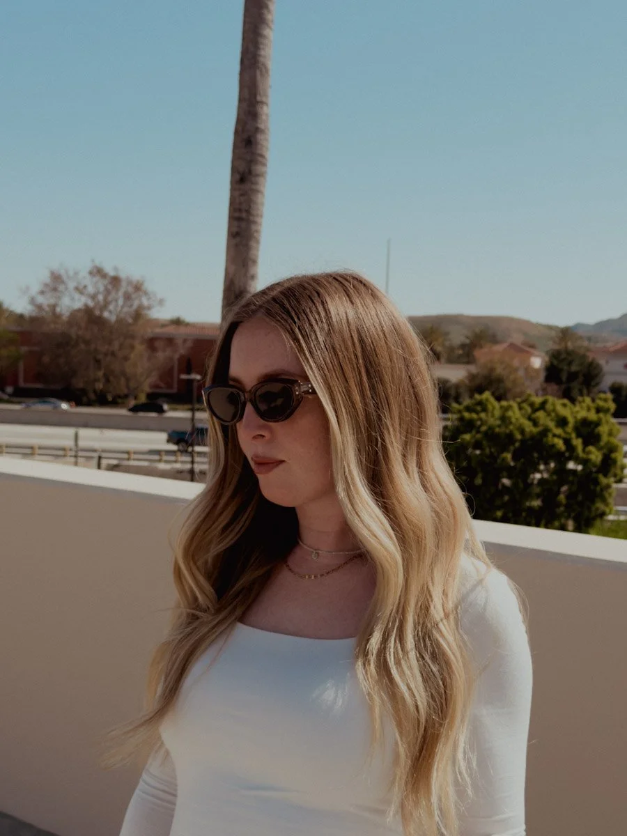 A young woman with long wavy blonde hair wearing sunglasses and a white top, standing outdoors with trees, buildings, and mountains in the background on a sunny day.