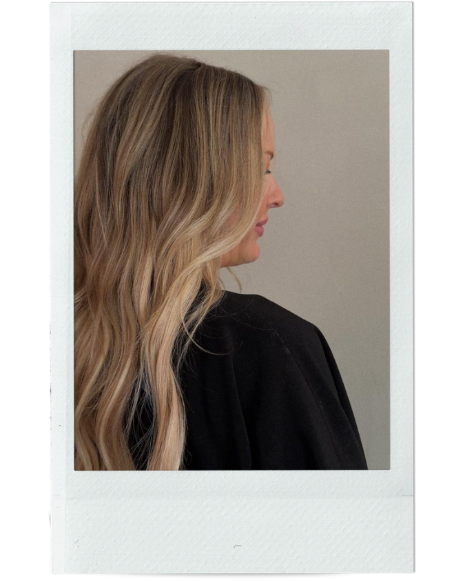 Side profile of a woman with long, wavy blonde hair wearing a black top, against a plain light-colored background.