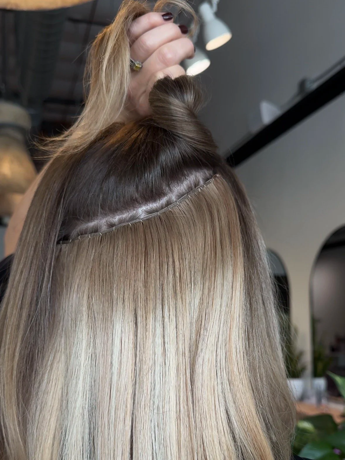 Hairdresser applying hair extensions or bonds to a woman's hair at a salon, with blonde hair and a section of hair being attached.