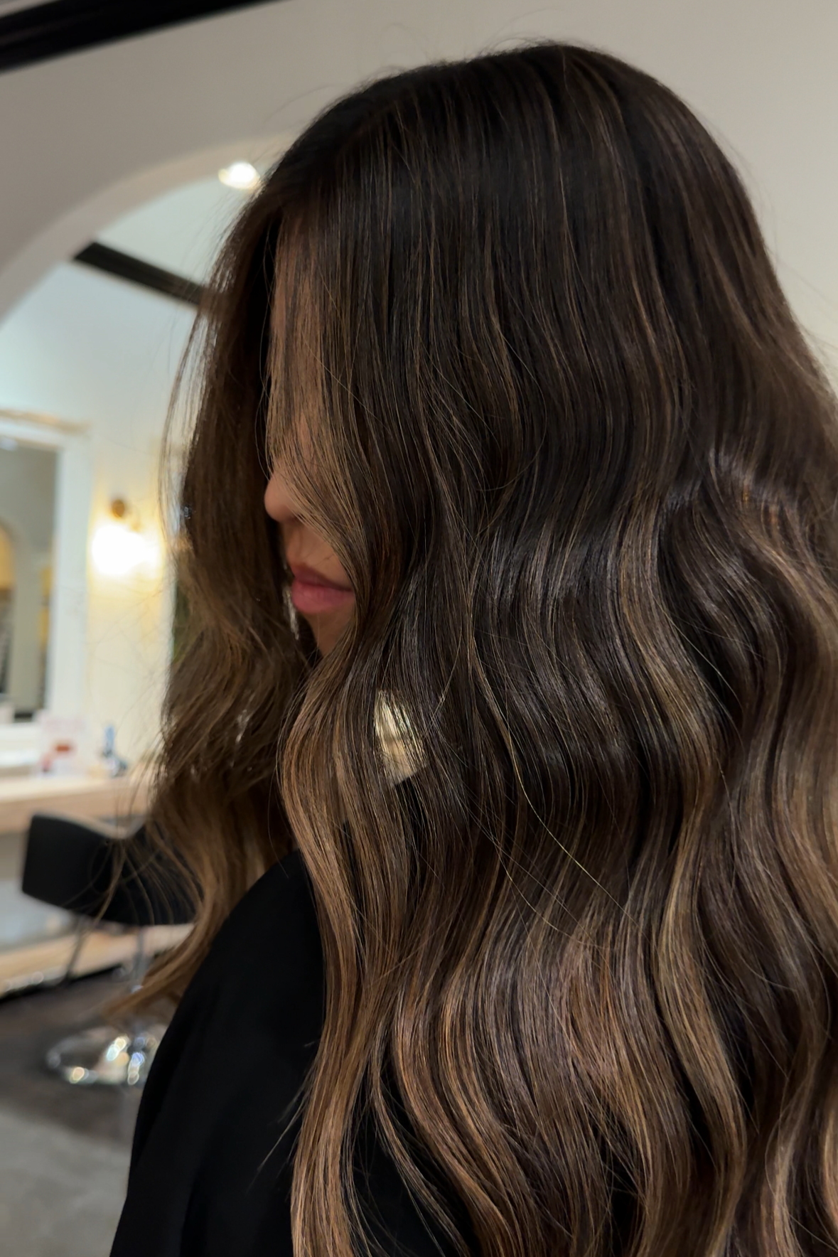 A woman with long, wavy, brown hair with highlights in a salon setting.