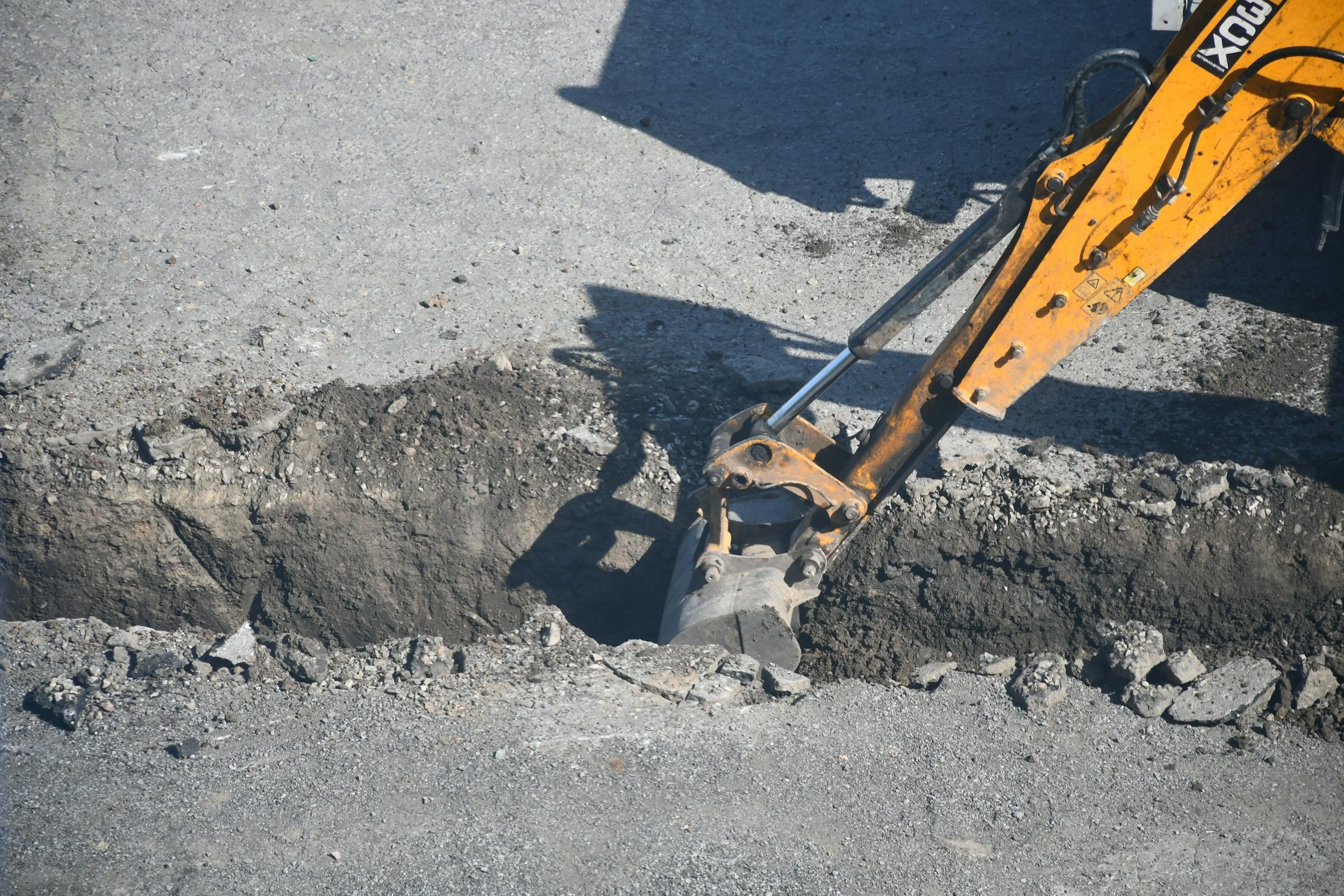 Close-up of a small excavator's arm digging a trench on a gravelly road.
