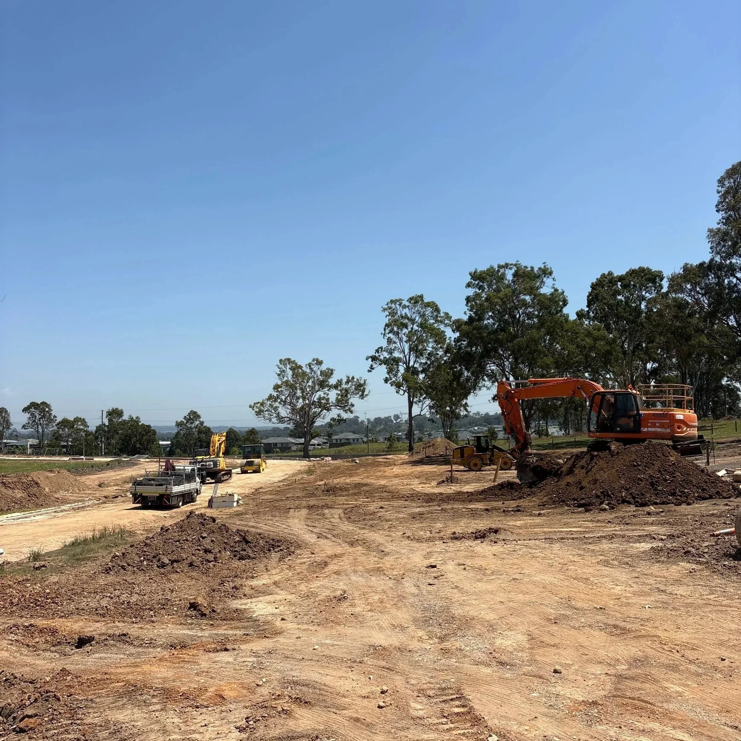 Construction site with excavator, bulldozer, trucks, and piles of dirt under a clear blue sky and trees in the background.