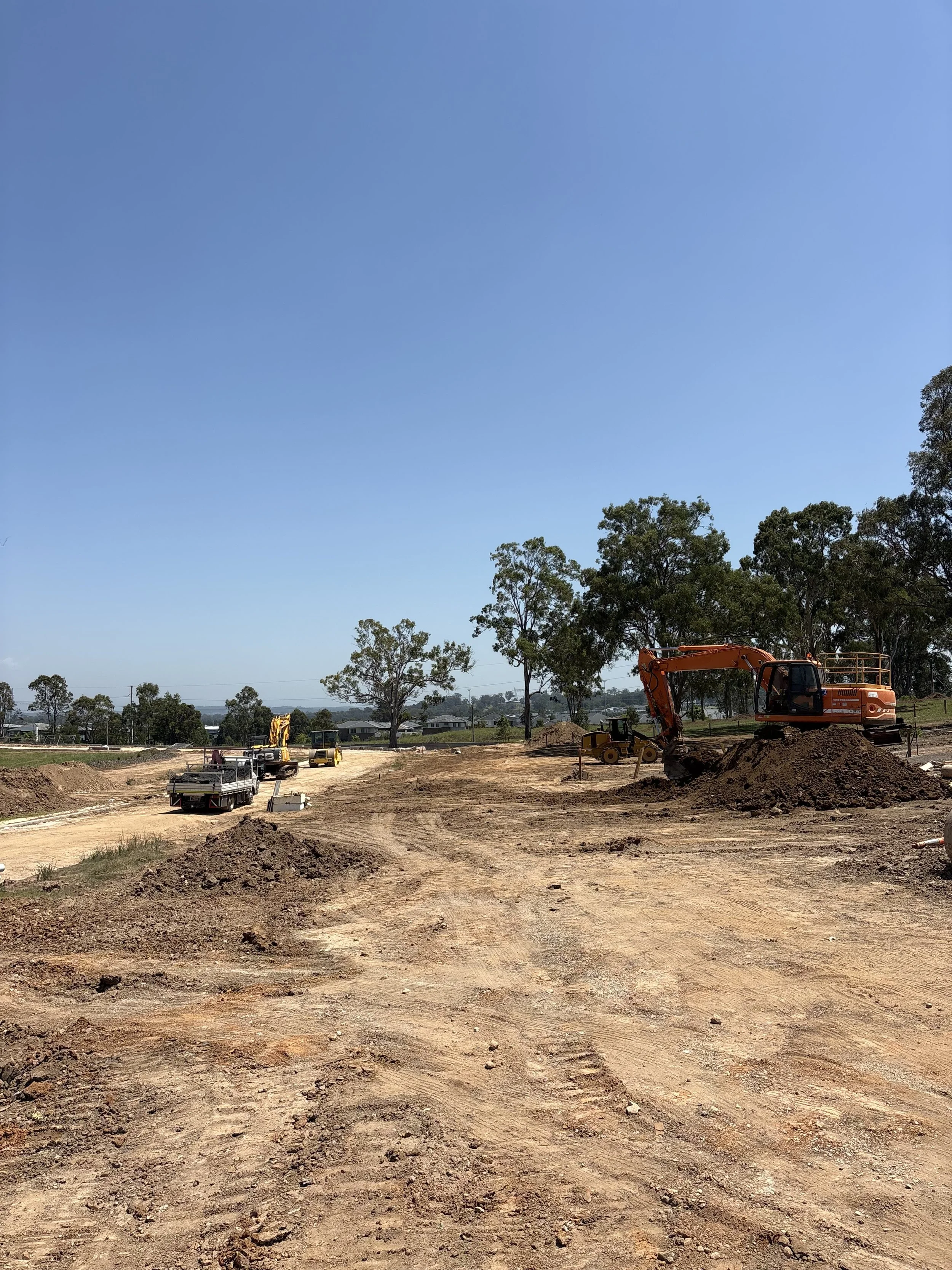 Construction site with orange excavator moving dirt and several smaller construction vehicles on dirt road under a clear blue sky, with trees in the background.