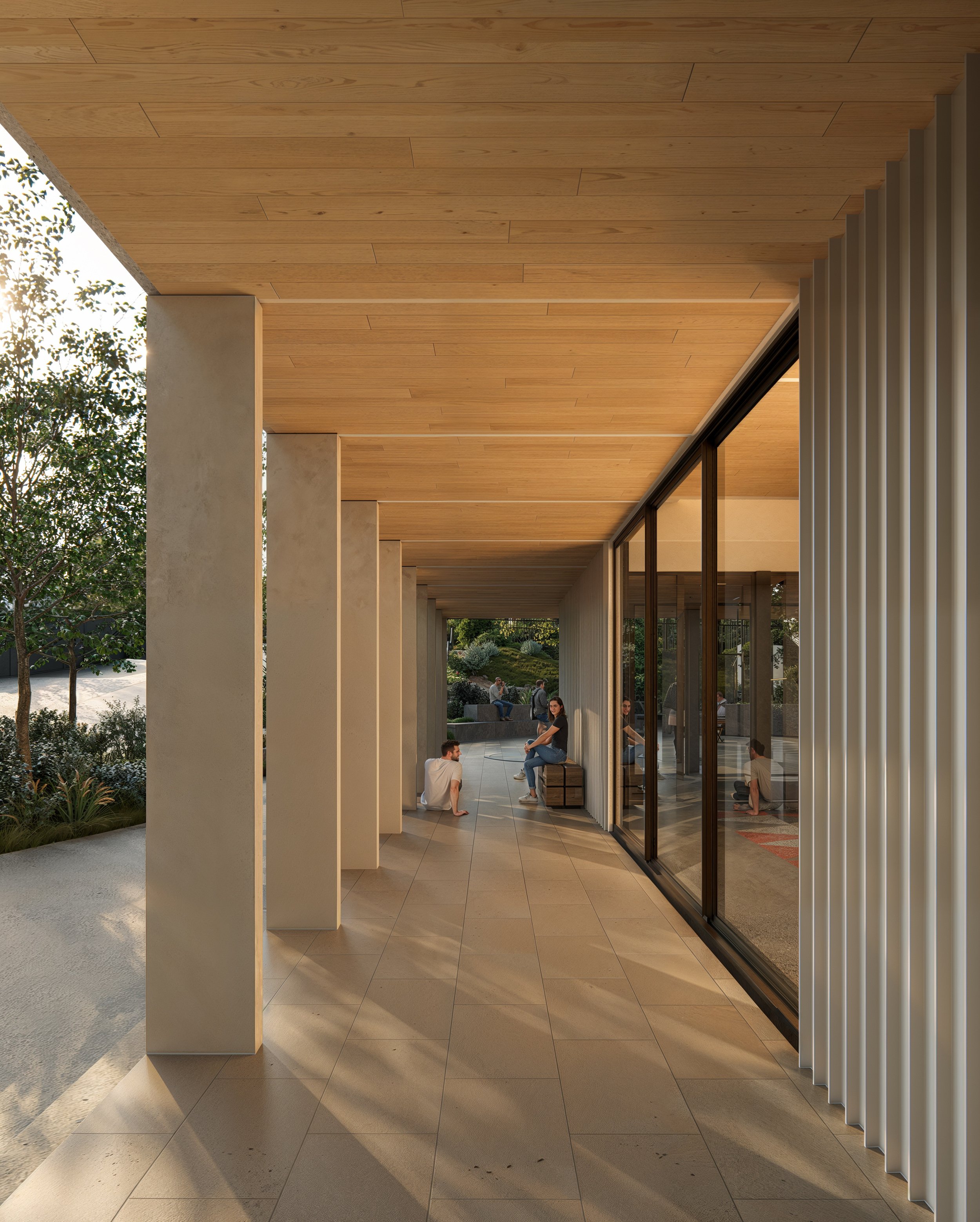 Modern building with a covered outdoor patio area featuring beige tiled flooring, concrete columns, a wooden ceiling, and large sliding glass doors. Several people are sitting and standing, engaging in conversations. The patio overlooks a landscaped 