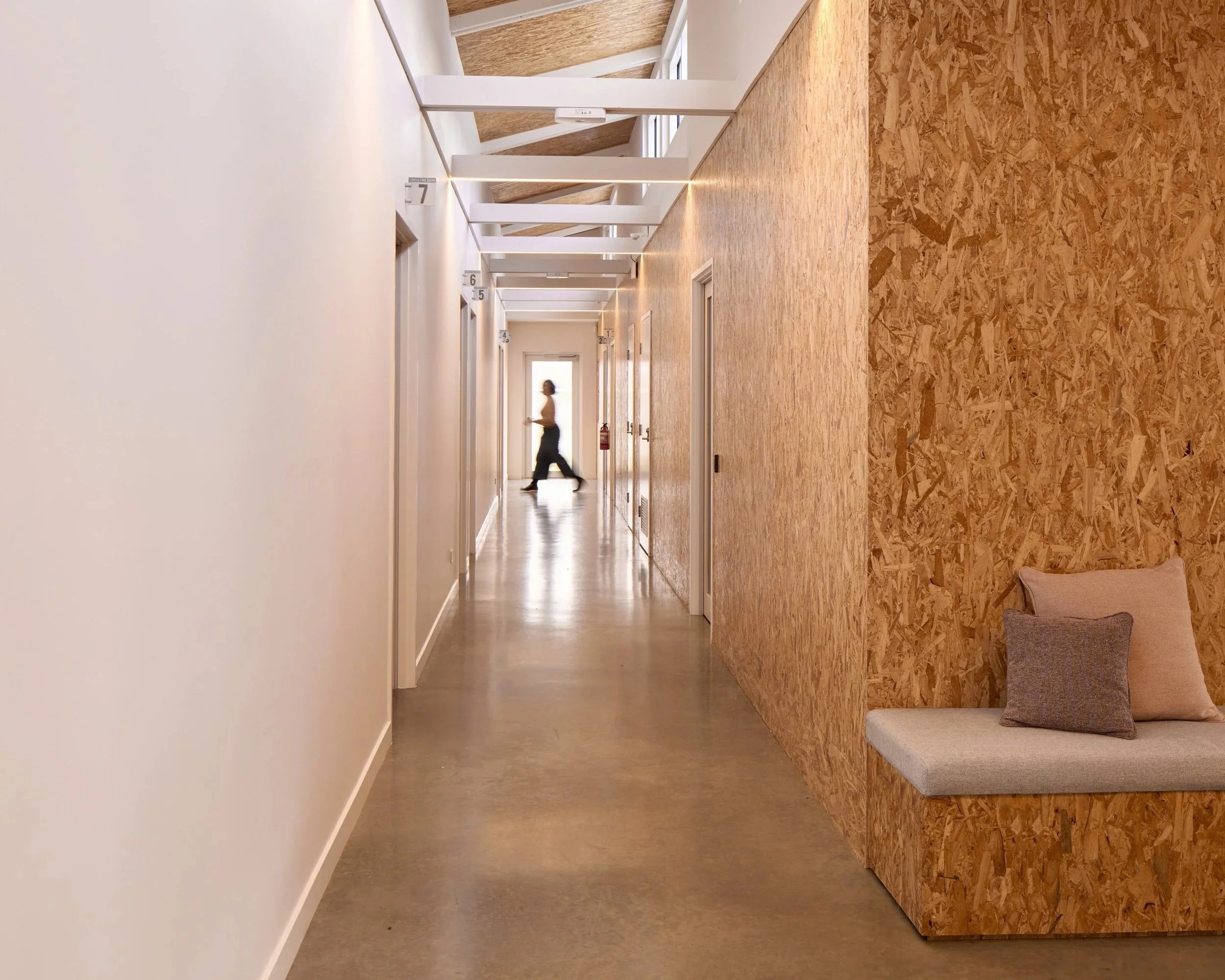 Interior view of a modern corridor with a wooden patterned wall, white ceiling, and flooring, containing benches and illuminated with ceiling lights, with a person walking in the background.