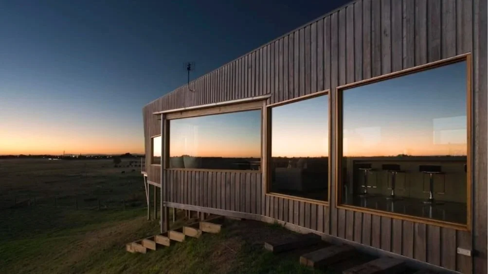 A modern wooden building featuring large windows reflecting a sunset over an open landscape, with a small set of wooden stairs leading to the entrance.