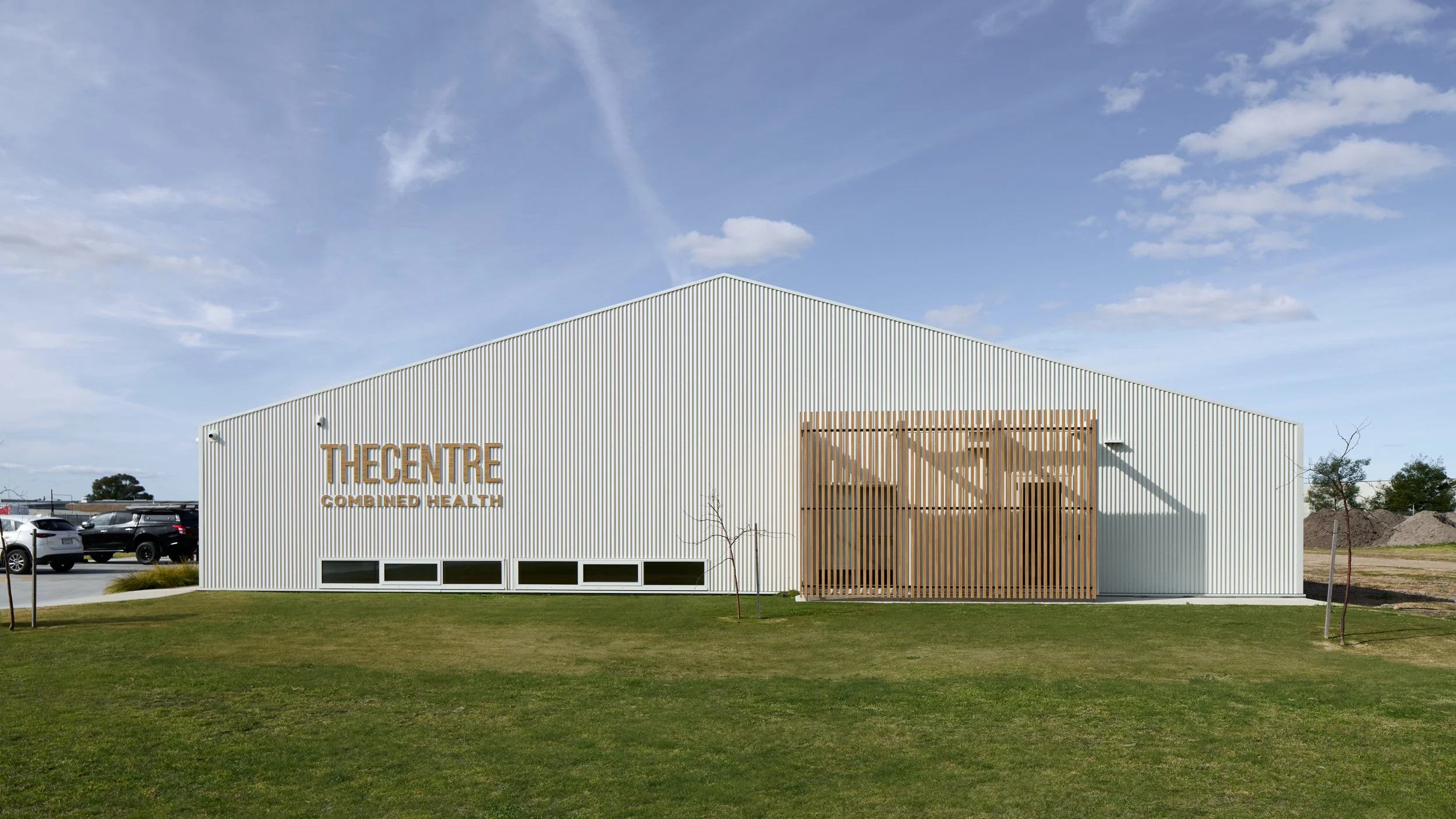 Exterior of a modern building with corrugated metal siding, wooden accents, and a sign that reads 'The Centre Combined Health.' There are cars parked nearby, small trees in the foreground, and a cloudy blue sky above.