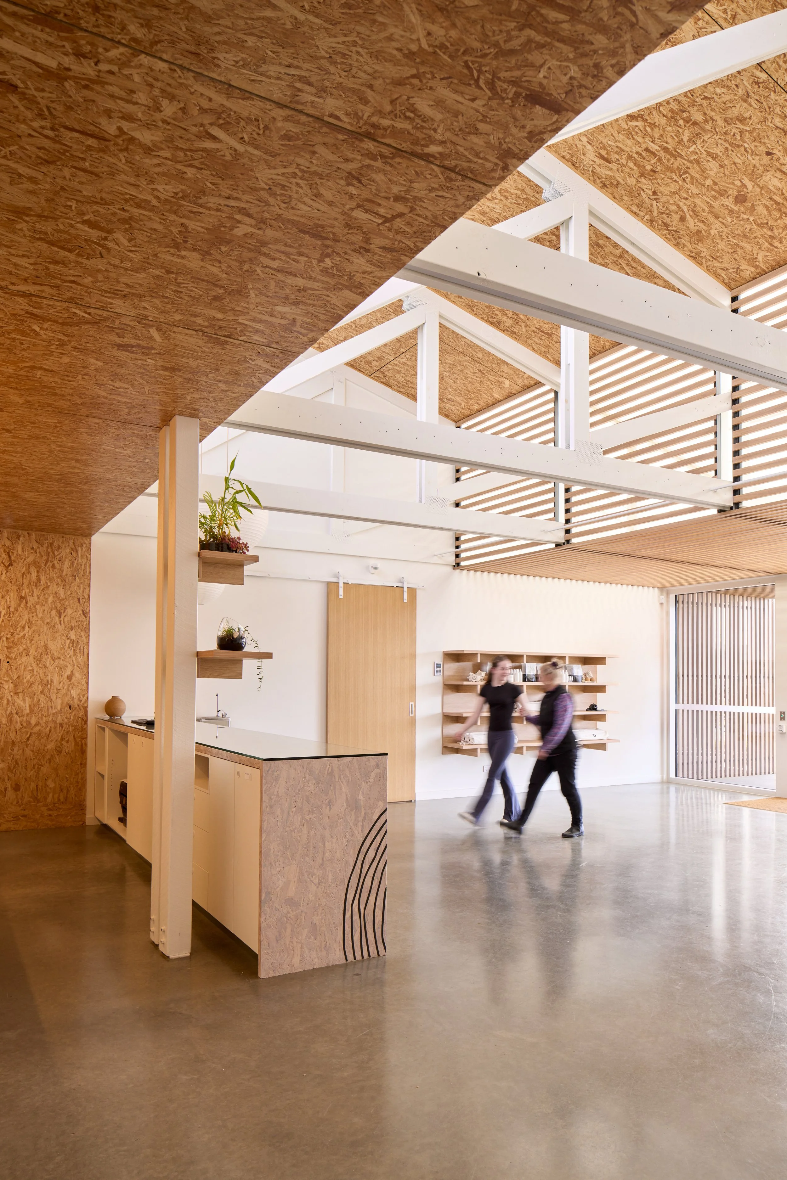 Modern indoor space with wooden ceiling, white structural beams, and a reception or kitchen area with shelves and plants. Two people are walking and talking in the open area, and there is a sliding door in the background.