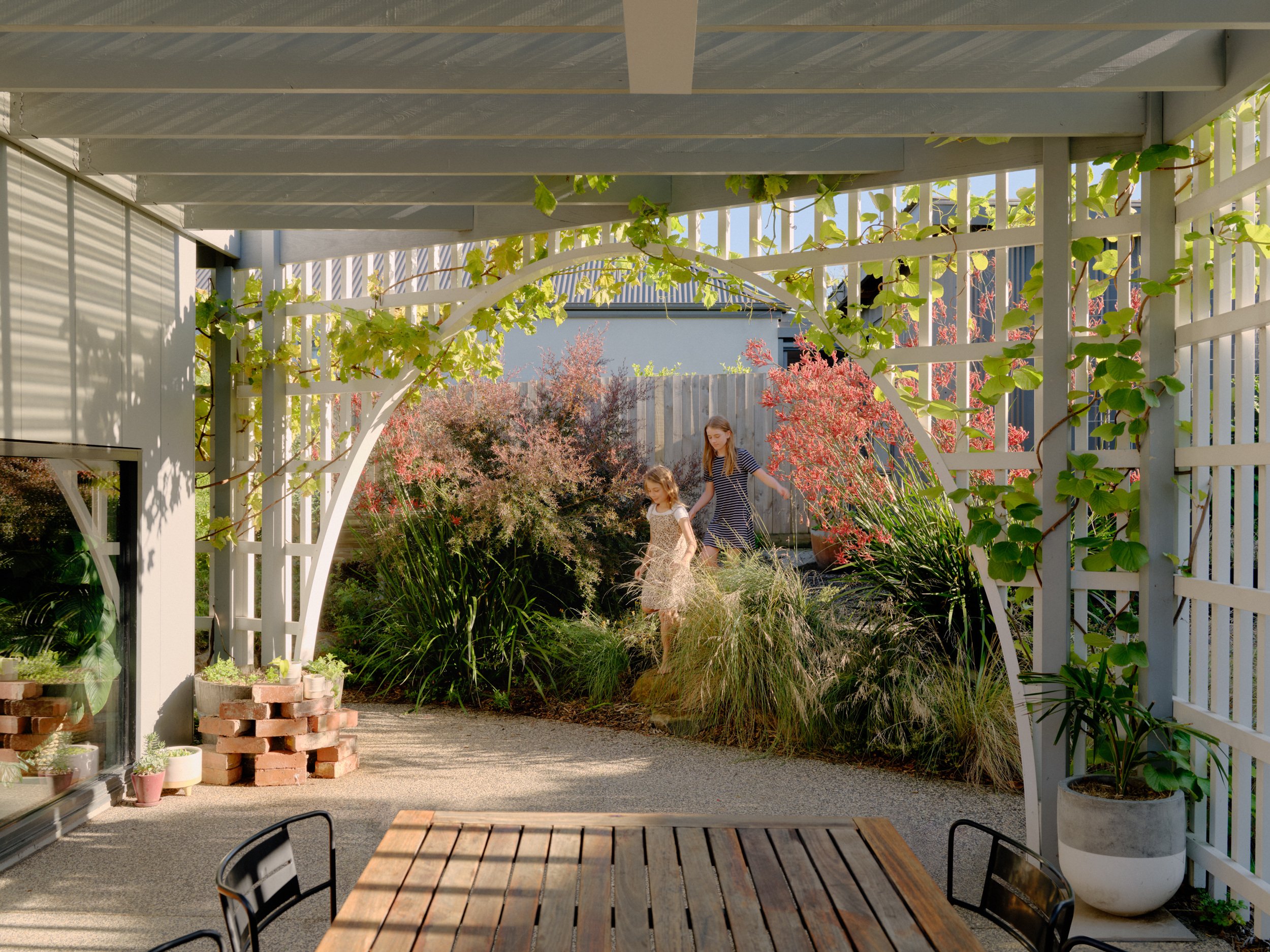 A cozy outdoor patio with a wooden table and black chairs, surrounded by lush greenery and flowering bushes, with a fence and a house in the background.