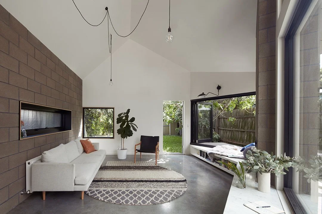 Modern living room with white sofa, black armchair, potted plant, abstract patterned rug, large windows, and minimalistic decor.