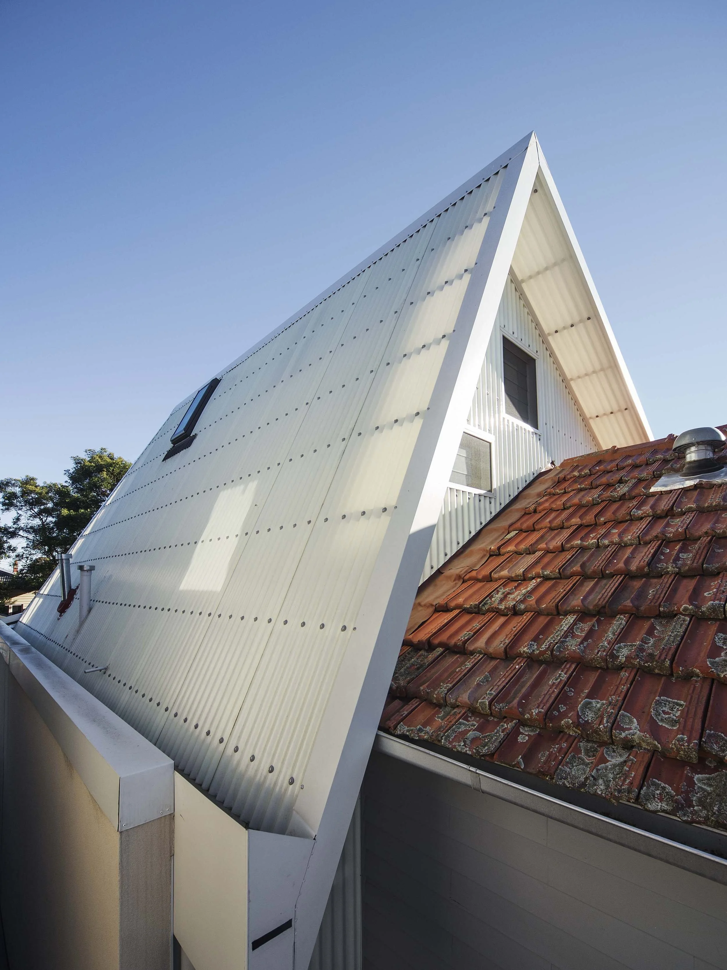 A modern house with a sharply angled white metal roof and a brick tiled roof section beneath it, against a clear blue sky.