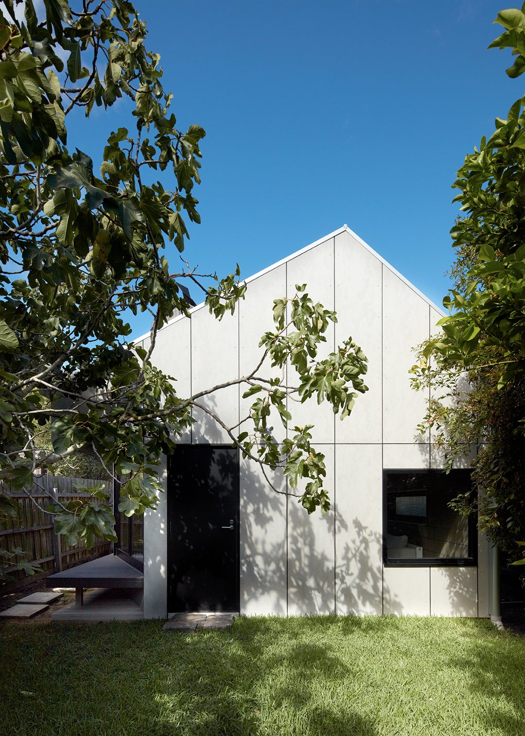 Modern white house with black door and window, surrounded by green trees and grass, under a clear blue sky.