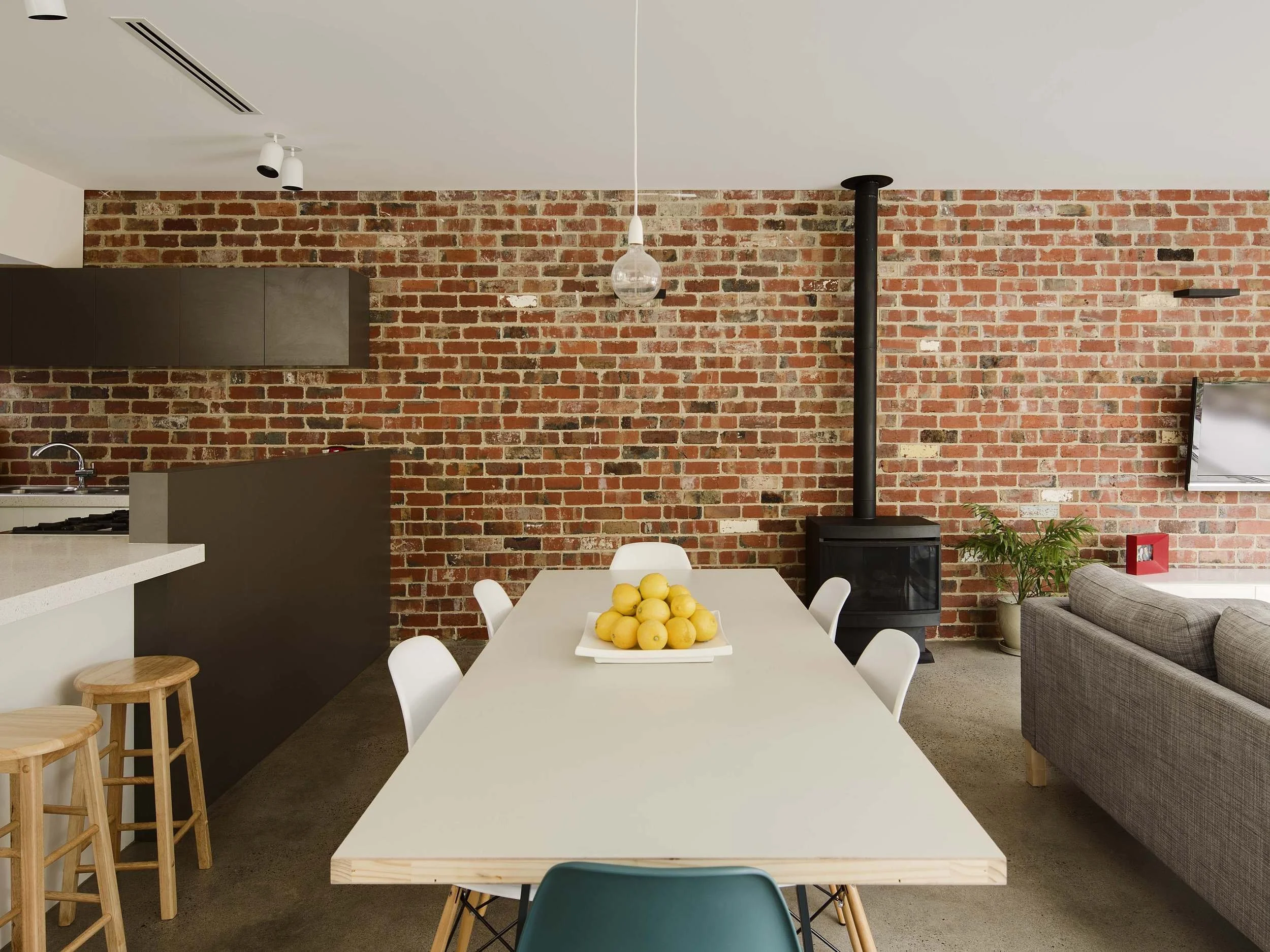 A modern kitchen and dining area with exposed red brick walls. The dining table has a bowl of yellow apples, surrounded by white chairs. There is a gray sofa and a small potted plant in the corner. The kitchen features dark cabinets, a sink, and a wh