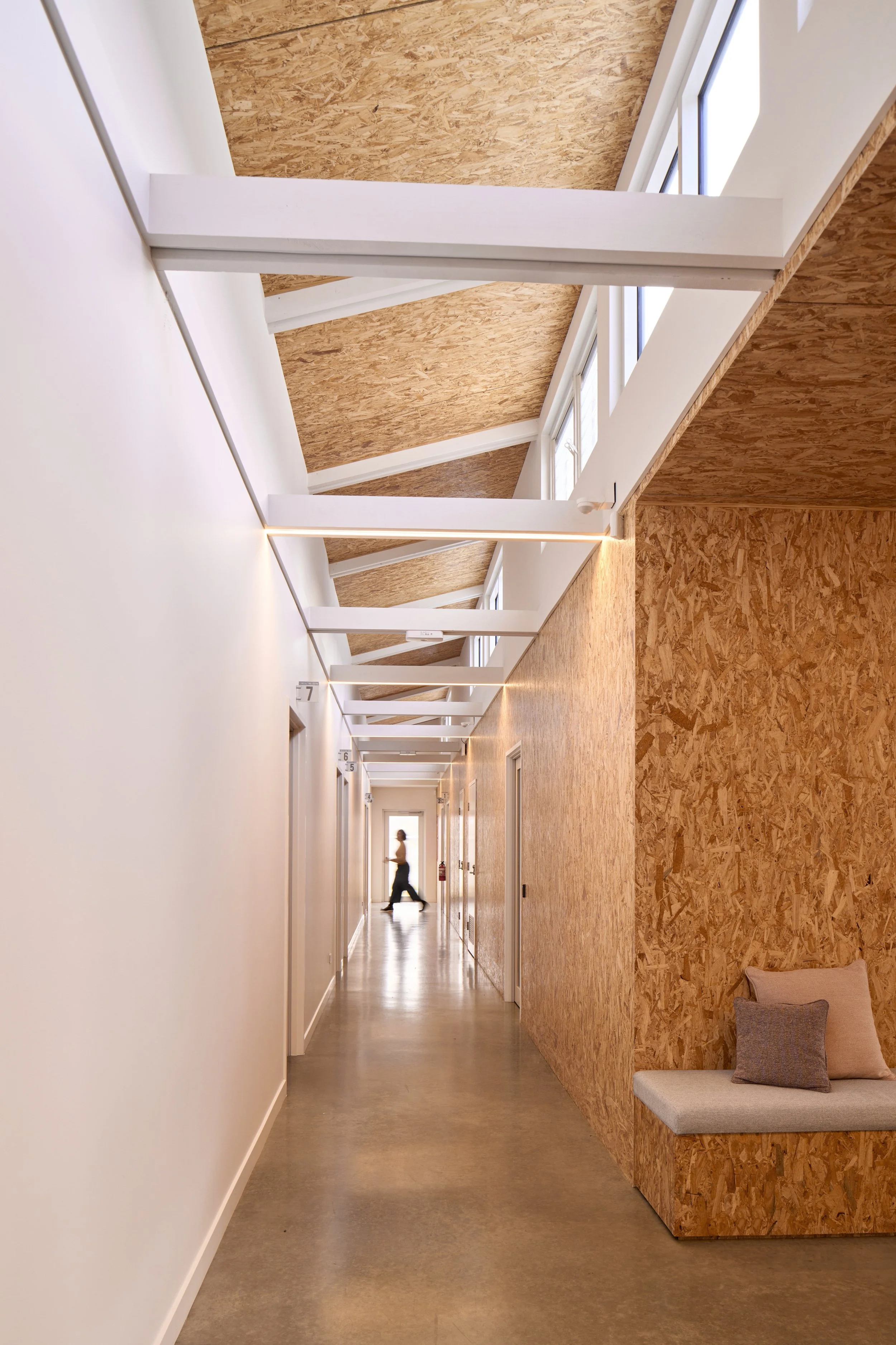 A modern, minimalistic hallway with white walls, a concrete floor, and a textured wood-paneled wall. The hallway has large windows on the ceiling allowing natural light to stream in. In the distance, there is a silhouette of a person walking.