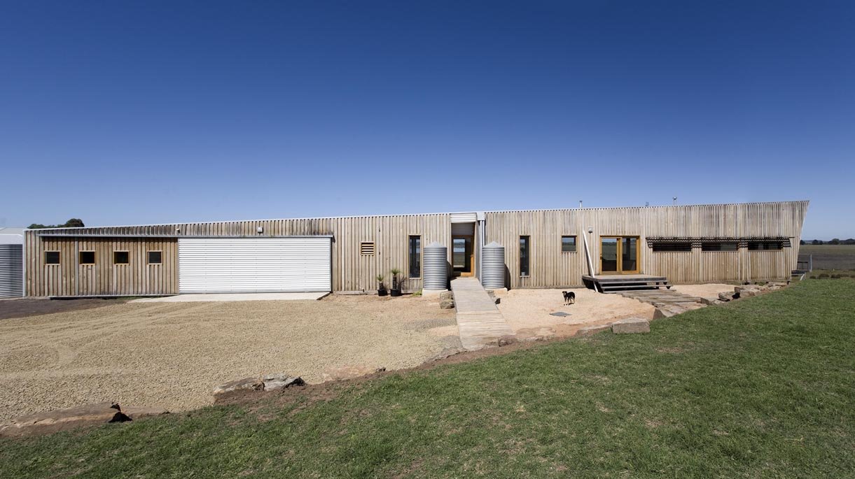 Modern house with wooden exterior, metal rainwater tanks near the entrance, small steps leading up to the door, and a dog standing on the sandy ground in front. Green grass patches in the foreground.