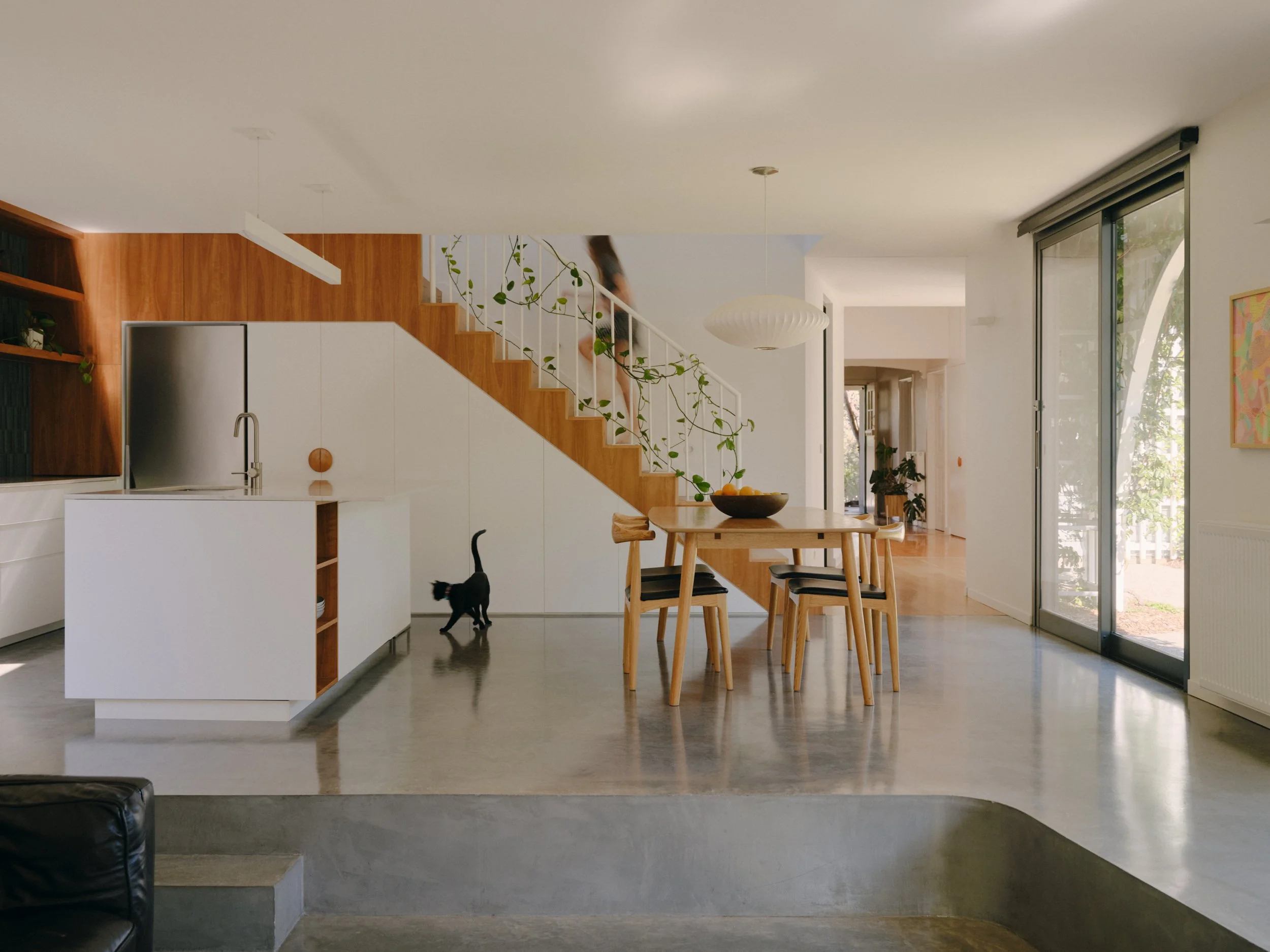 Modern open-plan kitchen and dining area with wooden staircase, green plants, round pendant light, large sliding glass doors, and black cat walking on the gray floor.