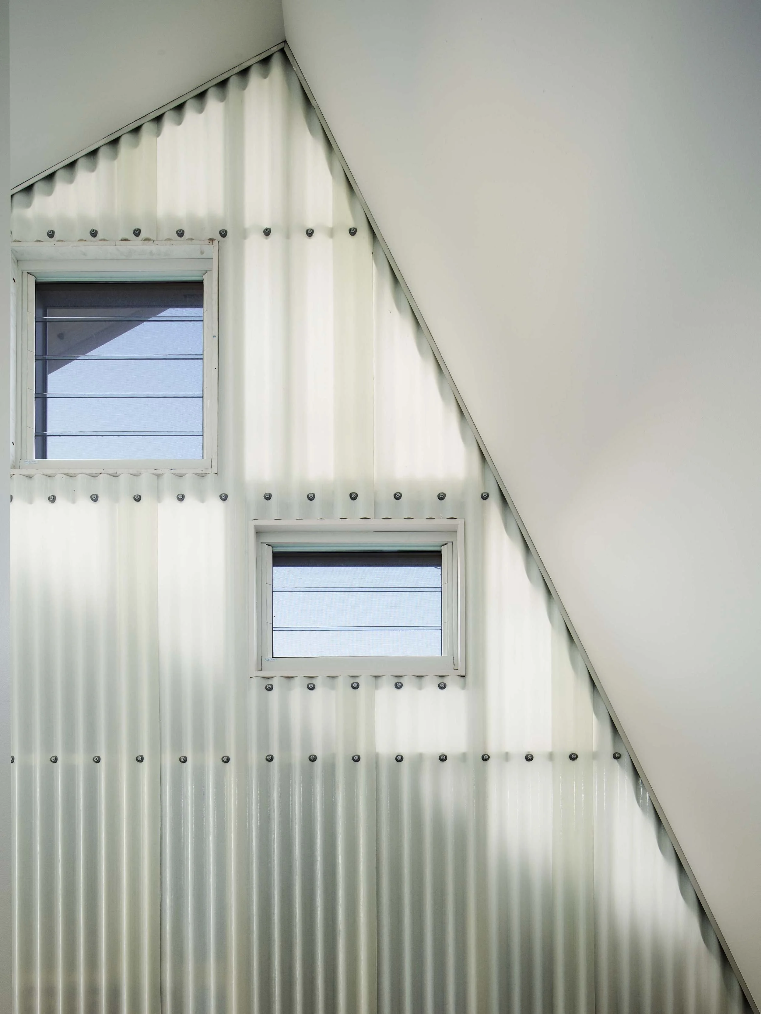 Interior view of a modern building with two small rectangular windows and a sloped ceiling. The windows have horizontal blinds and are surrounded by a metal wall with visible bolts.