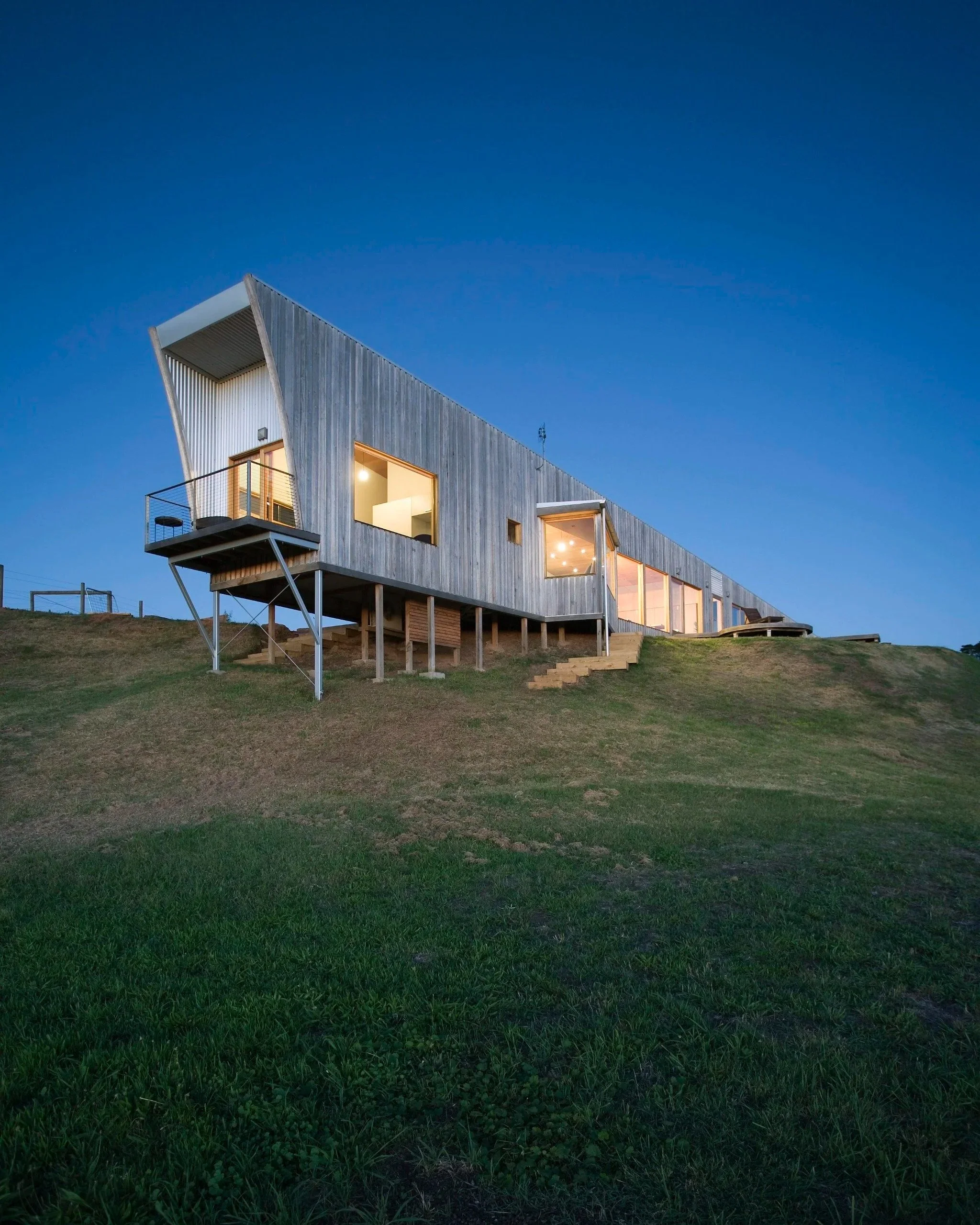 Modern house with large windows and a balcony, built on stilts on a grassy hillside during dusk.