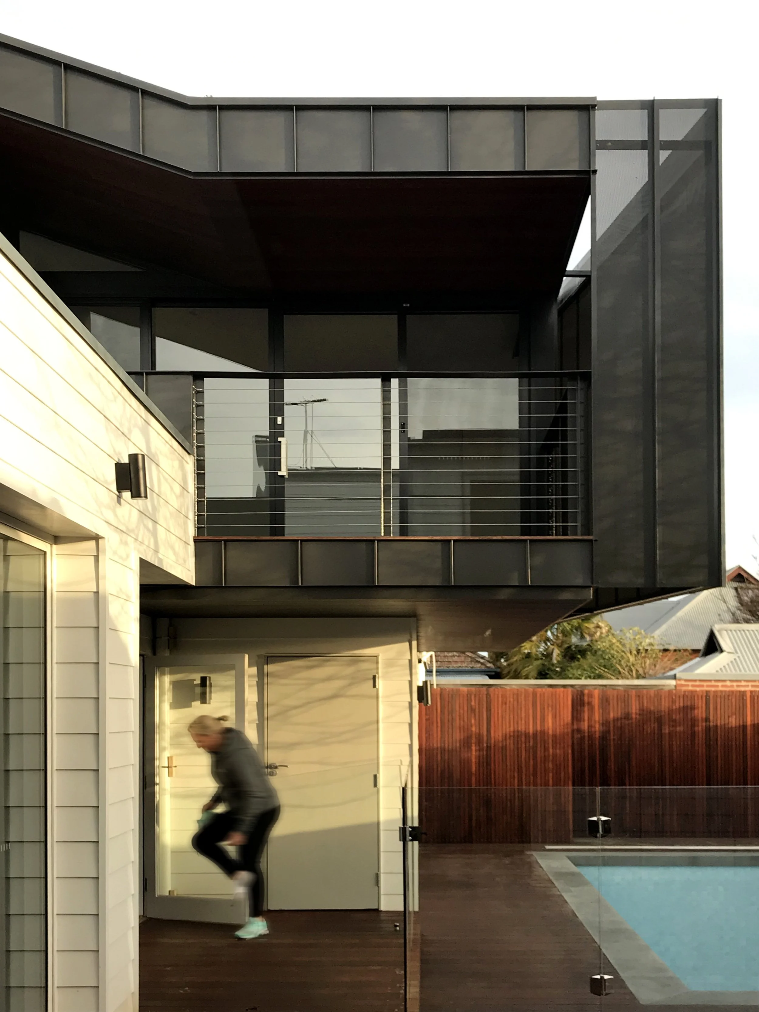Modern two-story house with black exterior, glass railing balcony, wooden deck, swimming pool, and sliding glass door, with a person walking on the deck.