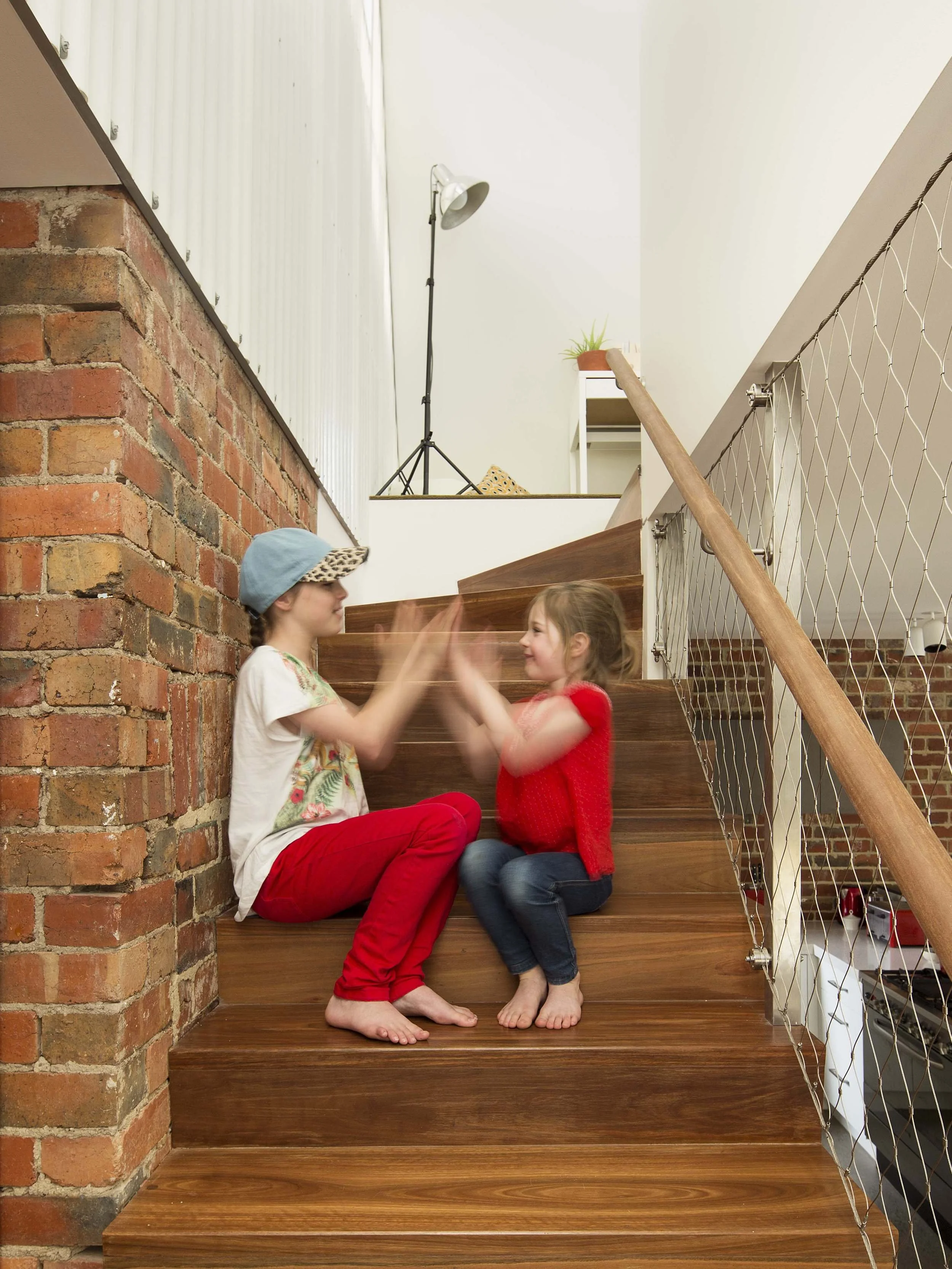 Two young girls sitting on wooden stairs, smiling and clapping hands, with one wearing a blue cap and the other in a red sweater, inside a home with exposed brick walls and metal wire railing.